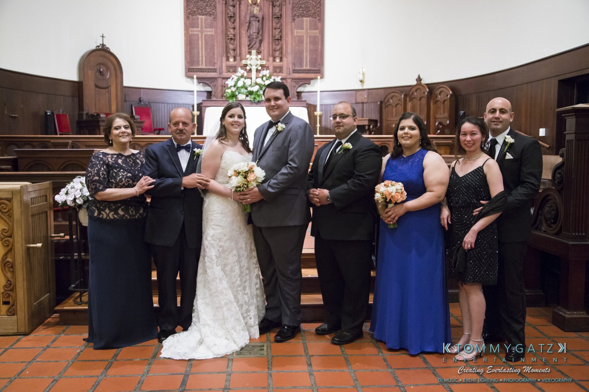 Jamie-and-Mark-Low-Res-Watermark-326 - TOMMY GATZ ENTERTAINMENT A wedding party poses for a photo in a church, including the bride, groom, and six others, all dressed in formal attire.