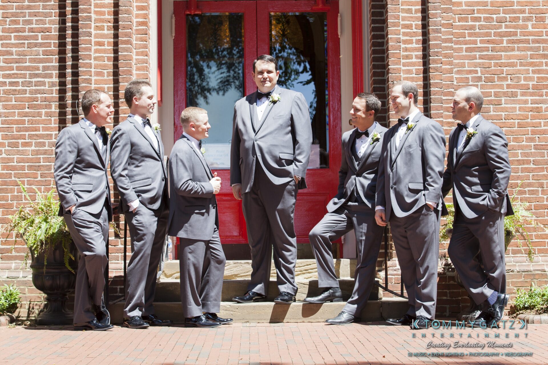 Jamie-and-Mark-Low-Res-Watermark-33 - TOMMY GATZ ENTERTAINMENT A group of seven men in gray suits stand and converse on steps in front of a building with red doors.