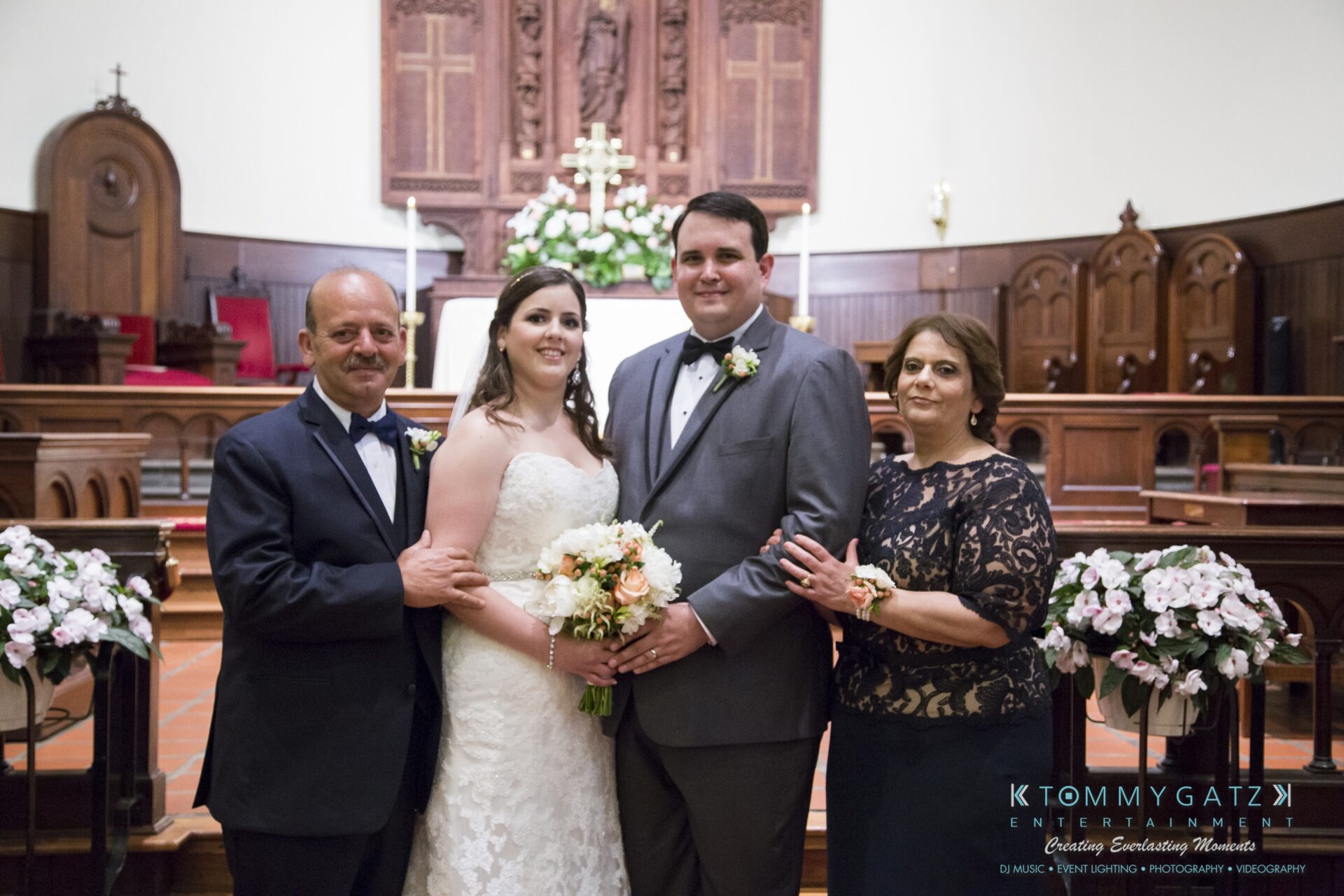 Jamie-and-Mark-Low-Res-Watermark-335 - TOMMY GATZ ENTERTAINMENT Bride and groom pose with two people in a church, with floral decorations and wooden pews in the background.