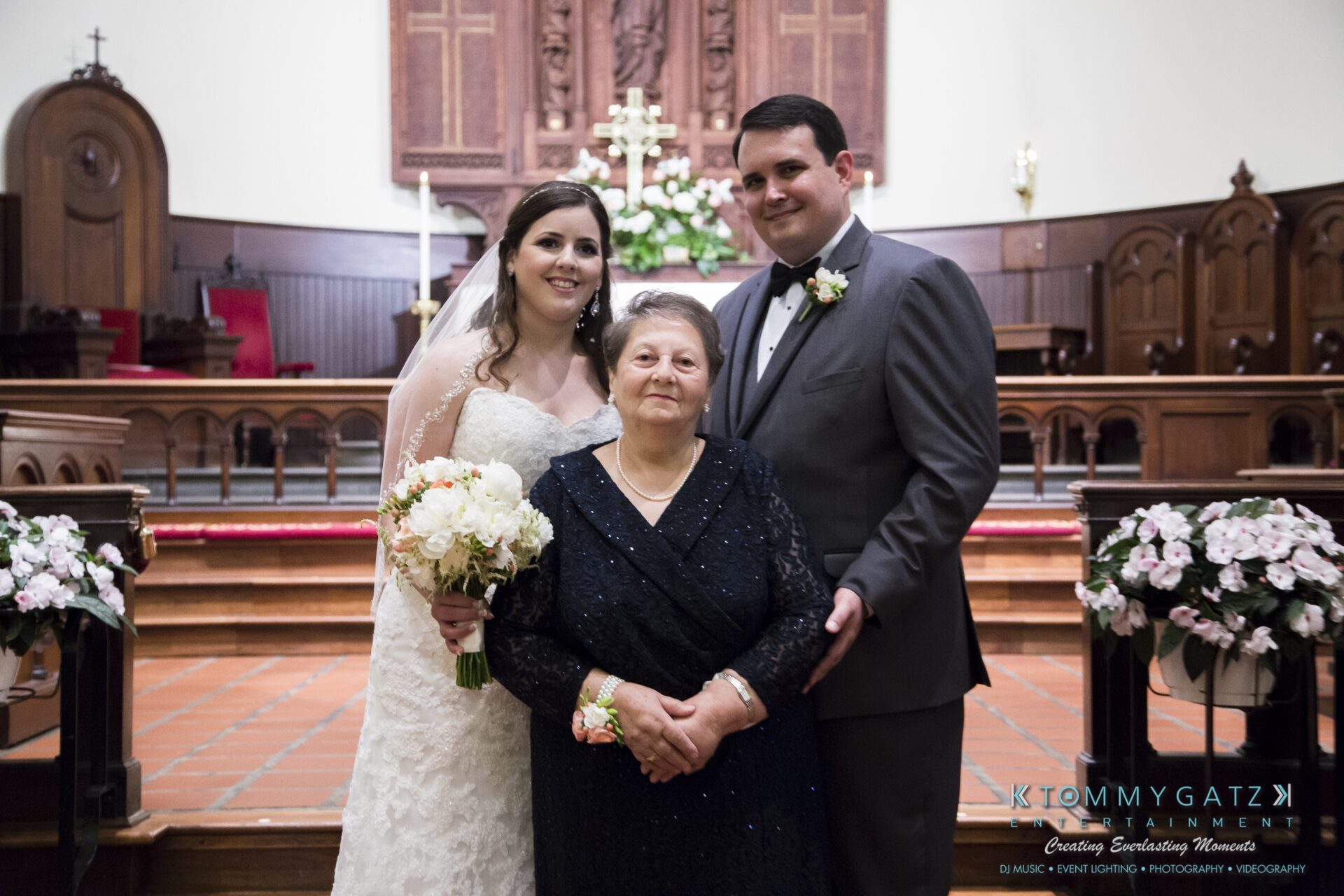 Jamie-and-Mark-Low-Res-Watermark-355 - TOMMY GATZ ENTERTAINMENT Bride and groom stand beside an older woman in a church setting, with flowers and wood paneling in the background.