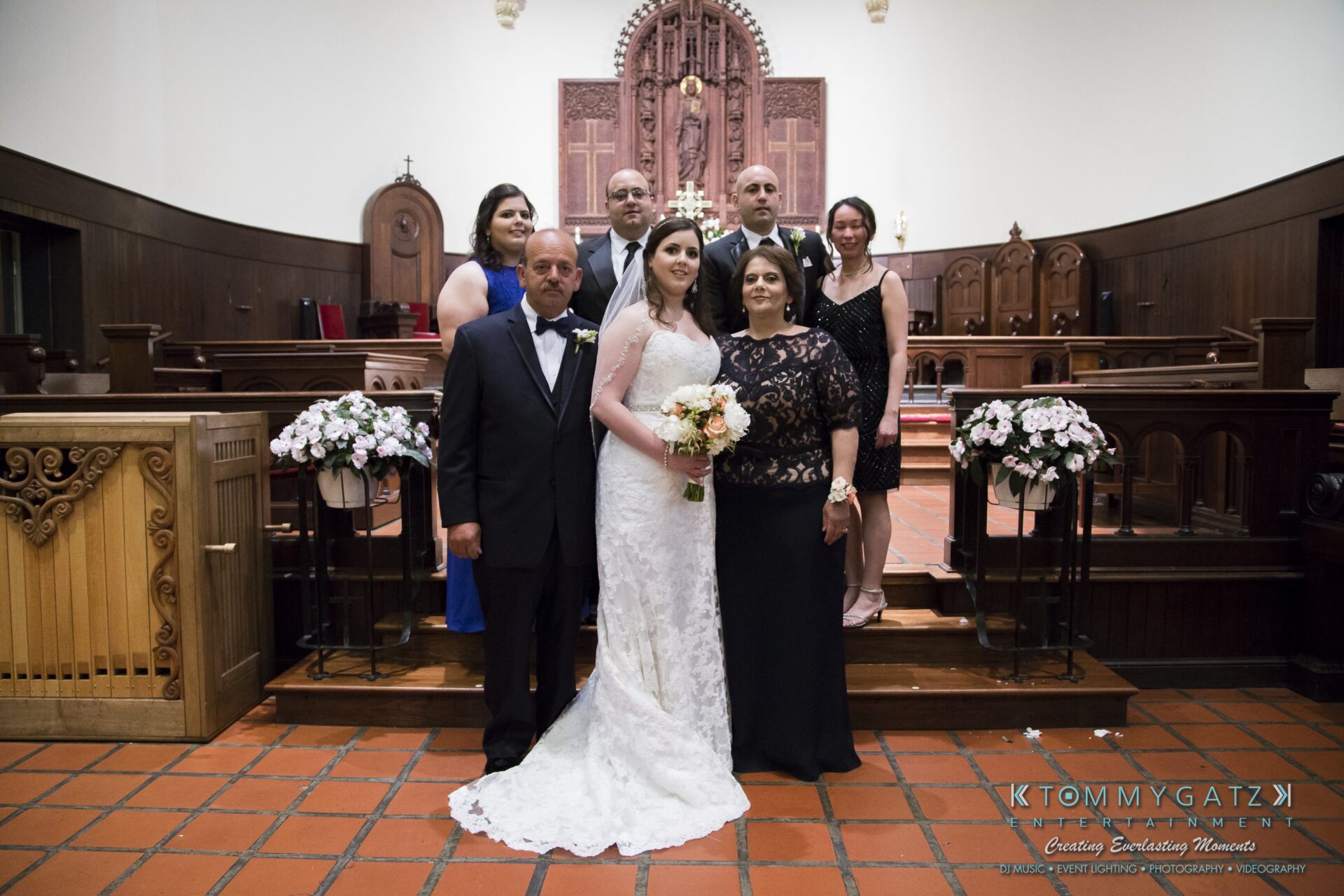 Jamie-and-Mark-Low-Res-Watermark-371 - TOMMY GATZ ENTERTAINMENT A bride and groom pose with their family in a church setting. The bride holds a bouquet and wears a lace gown. The family members are dressed formally, standing on terracotta tiles.