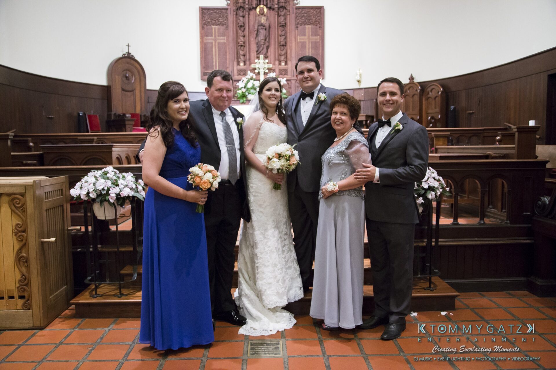 Jamie-and-Mark-Low-Res-Watermark-379 - TOMMY GATZ ENTERTAINMENT A wedding party poses in a church, featuring the bride and groom in the center, flanked by four other people. The group stands in front of a wooden altar with floral arrangements.