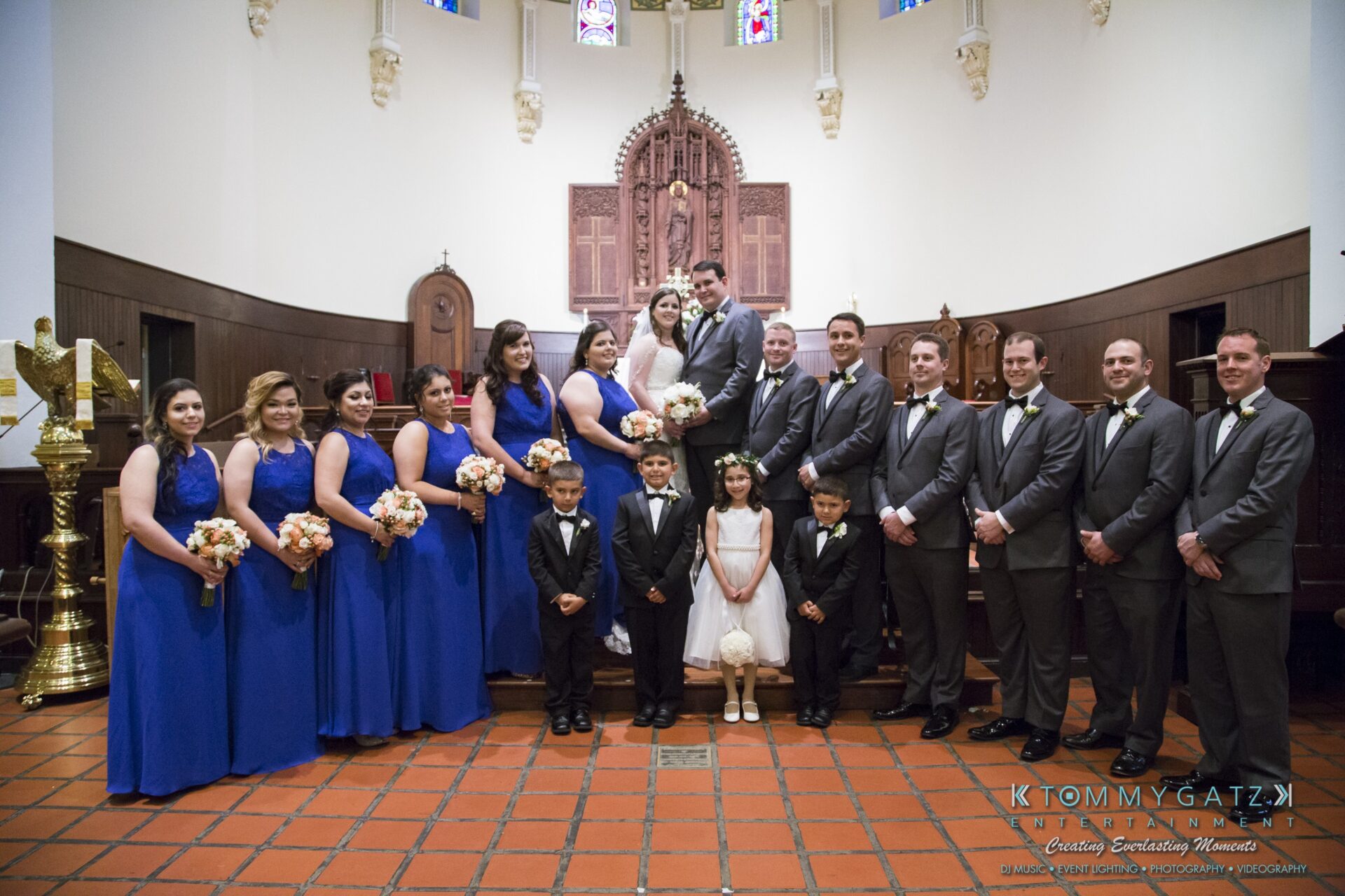 Jamie-and-Mark-Low-Res-Watermark-393 - TOMMY GATZ ENTERTAINMENT A wedding party poses inside a church. The group includes the bride, groom, bridesmaids in blue dresses, groomsmen in suits, and three children dressed formally.