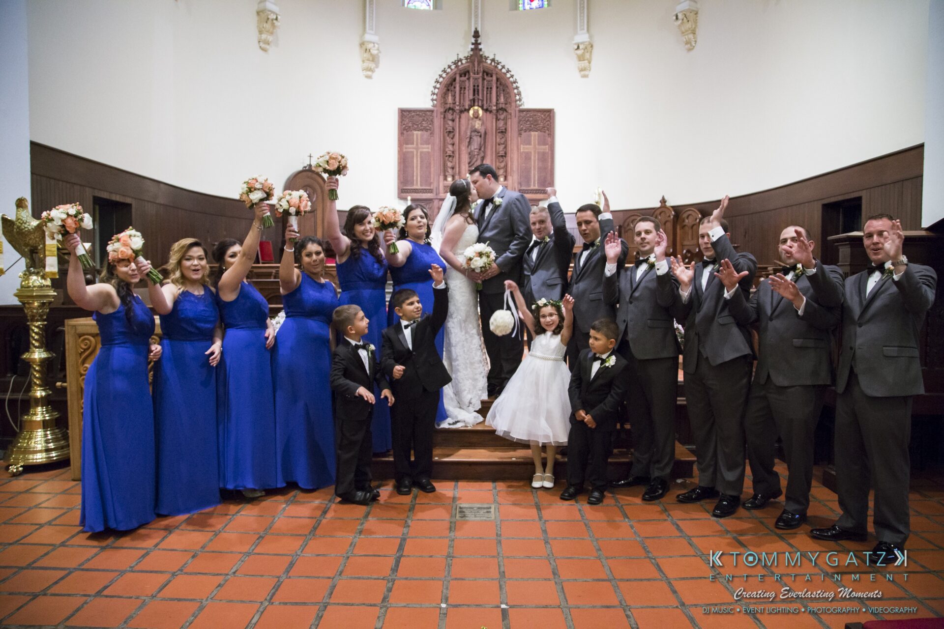 Jamie-and-Mark-Low-Res-Watermark-407 - TOMMY GATZ ENTERTAINMENT A wedding party celebrates on a church altar. The couple kisses in the center, surrounded by the bridal party in blue dresses and gray suits, holding bouquets. Children stand at the front.