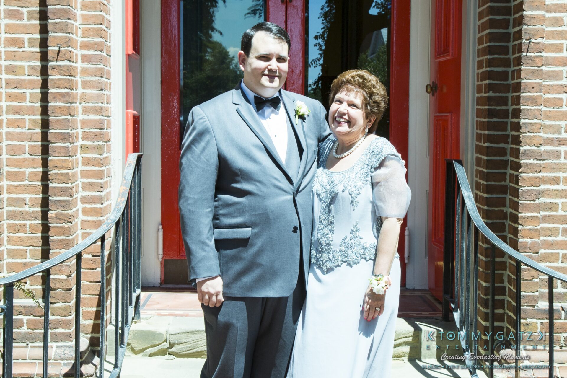 Jamie-and-Mark-Low-Res-Watermark-41 - TOMMY GATZ ENTERTAINMENT A man in a gray suit stands next to a smiling woman in a gray dress outside a brick building with red doors.