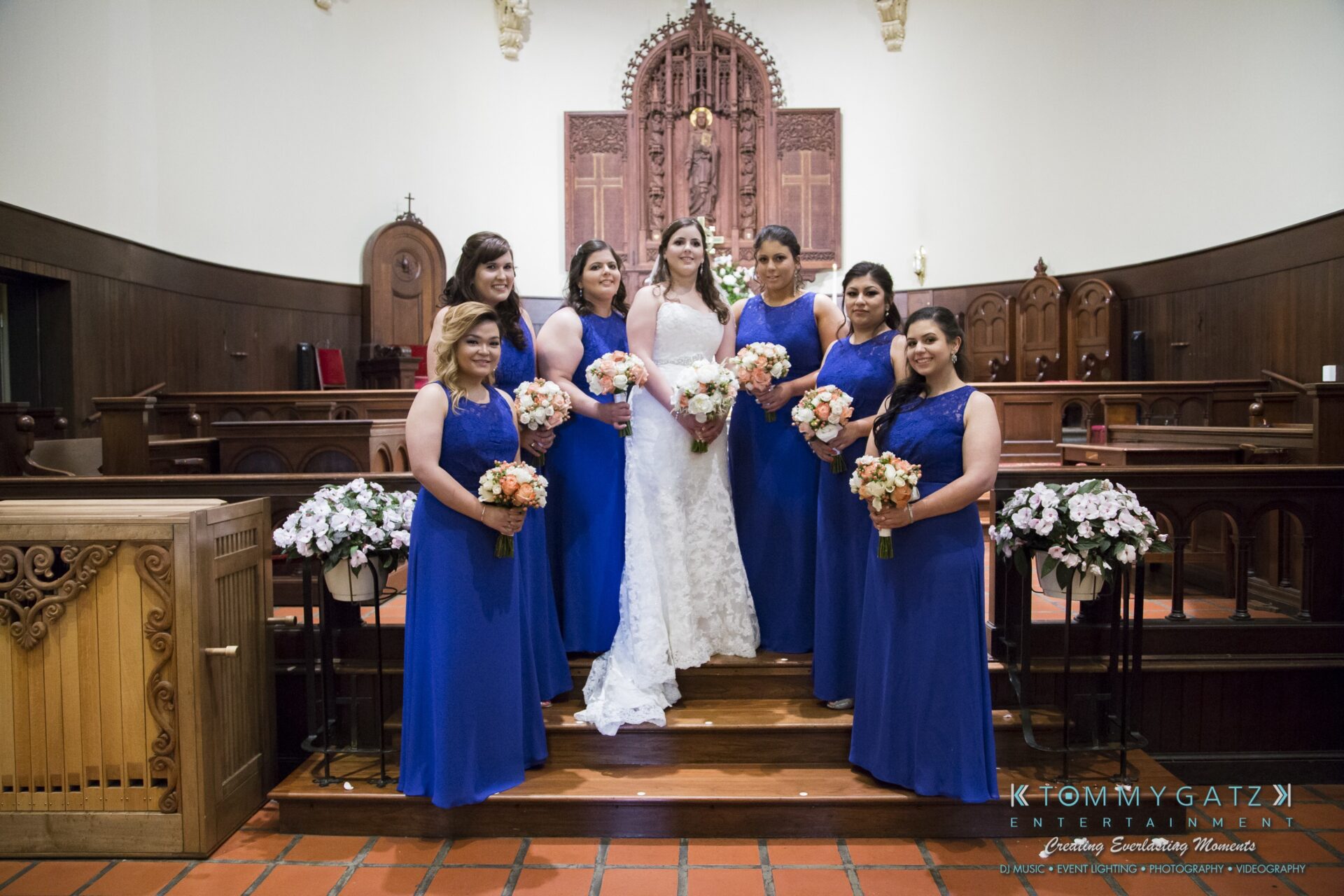 Jamie-and-Mark-Low-Res-Watermark-426 - TOMMY GATZ ENTERTAINMENT A bride in a white dress stands with six bridesmaids in blue dresses, each holding a bouquet, in a church with wooden pews and an ornate altar.