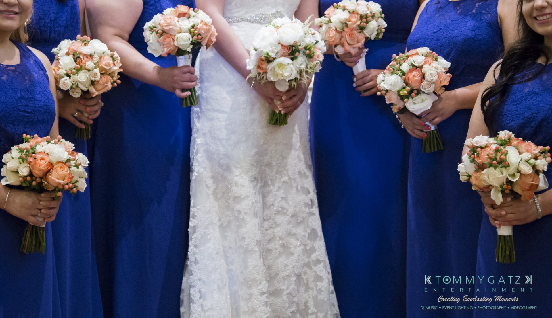 Jamie-and-Mark-Low-Res-Watermark-427 - TOMMY GATZ ENTERTAINMENT Bridesmaids in blue dresses holding bouquets of white and peach flowers surround a bride in a white gown.