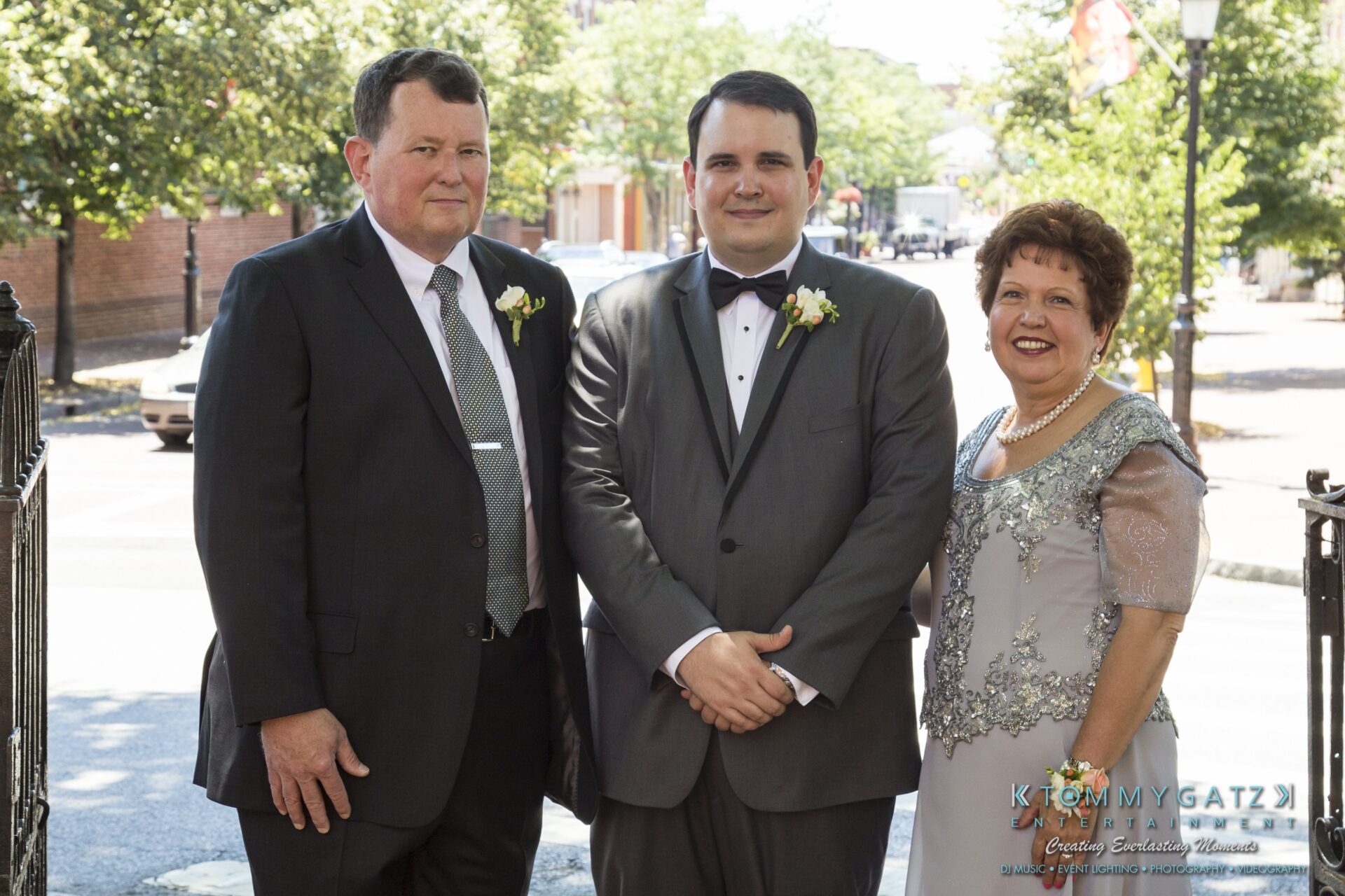 Jamie-and-Mark-Low-Res-Watermark-43 - TOMMY GATZ ENTERTAINMENT Three people dressed in formal attire, standing outdoors near a gate on a sunny day. They appear to be at a formal event or occasion, such as a wedding.
