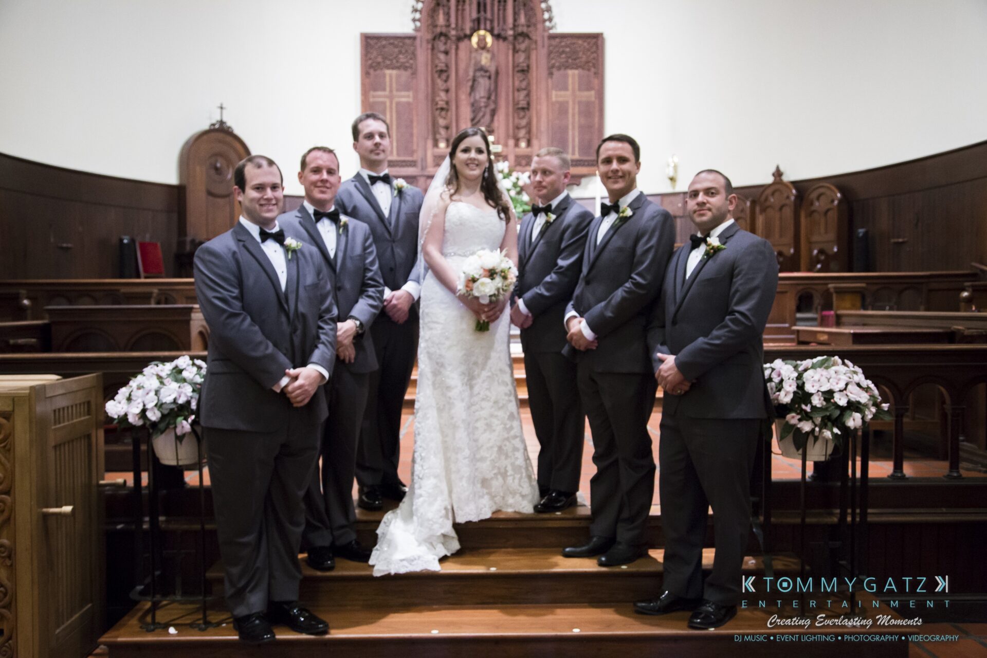Jamie-and-Mark-Low-Res-Watermark-431 - TOMMY GATZ ENTERTAINMENT A bride and seven groomsmen pose in a church, dressed in formal attire. The group is smiling and standing in front of an ornate wooden altar.