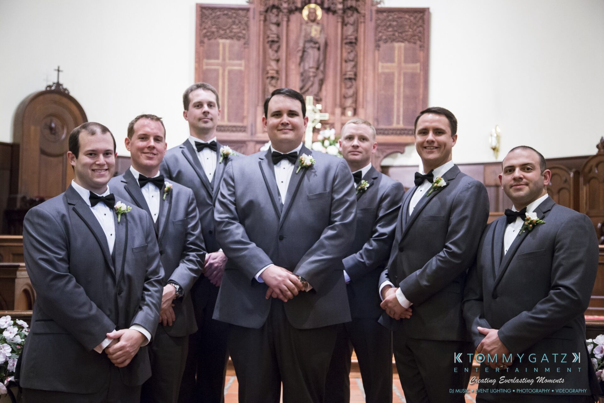 Jamie-and-Mark-Low-Res-Watermark-437 - TOMMY GATZ ENTERTAINMENT Seven groomsmen in tuxedos pose in a church setting with floral arrangements and wood paneling in the background.