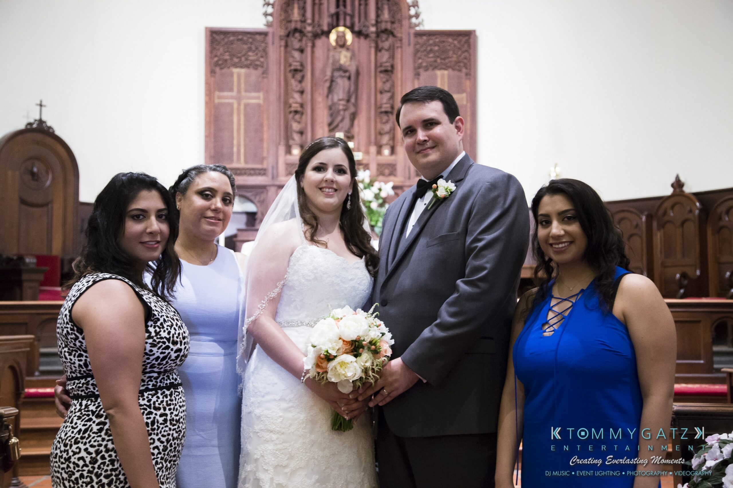 Jamie-and-Mark-Low-Res-Watermark-442 - TOMMY GATZ ENTERTAINMENT A bride and groom pose with three women in a church setting. The bride holds a bouquet, and everyone is dressed formally.