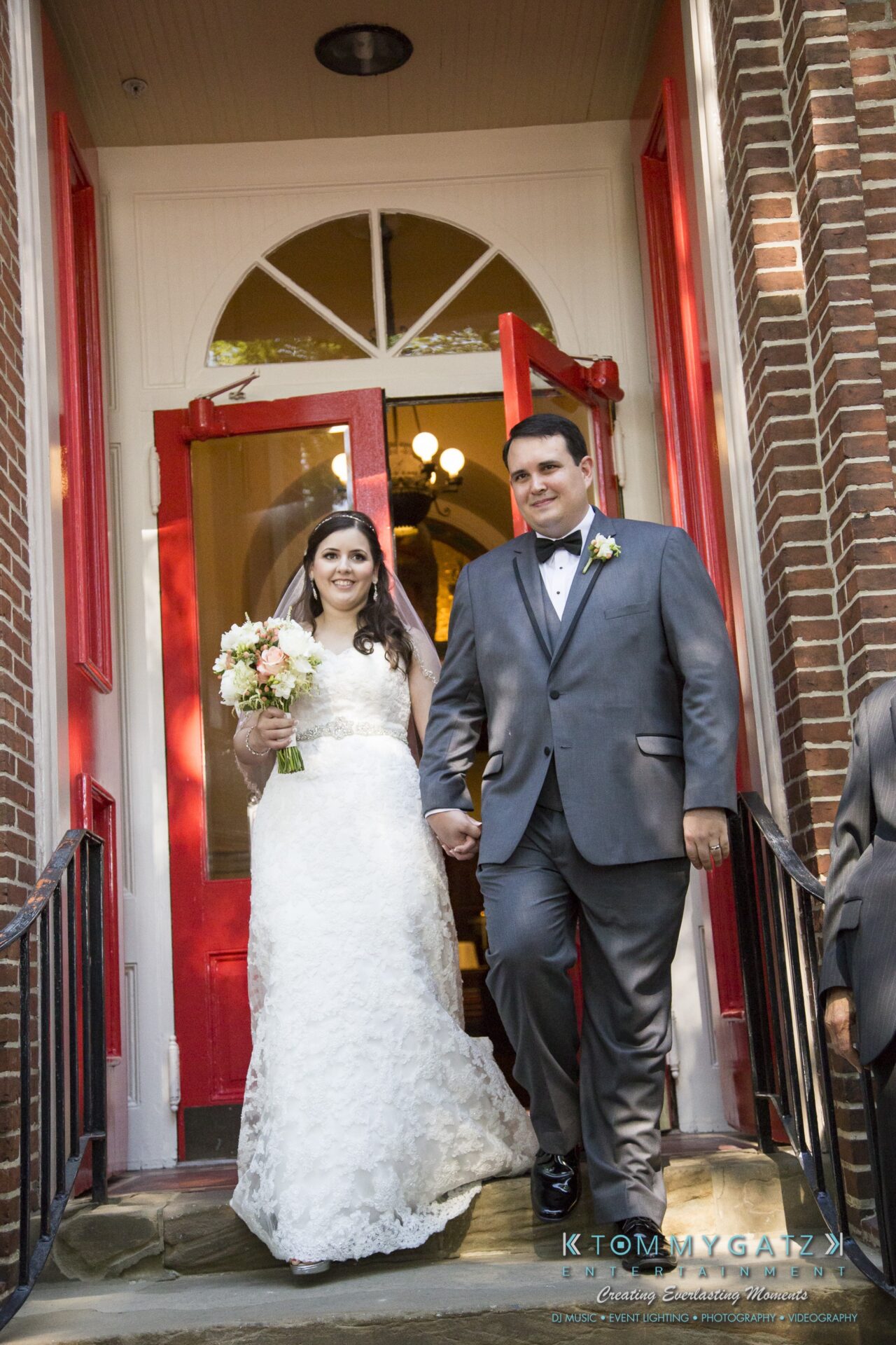Jamie-and-Mark-Low-Res-Watermark-446 - TOMMY GATZ ENTERTAINMENT Bride holding a bouquet and groom smiling as they walk down steps, framed by a red door and brick walls.
