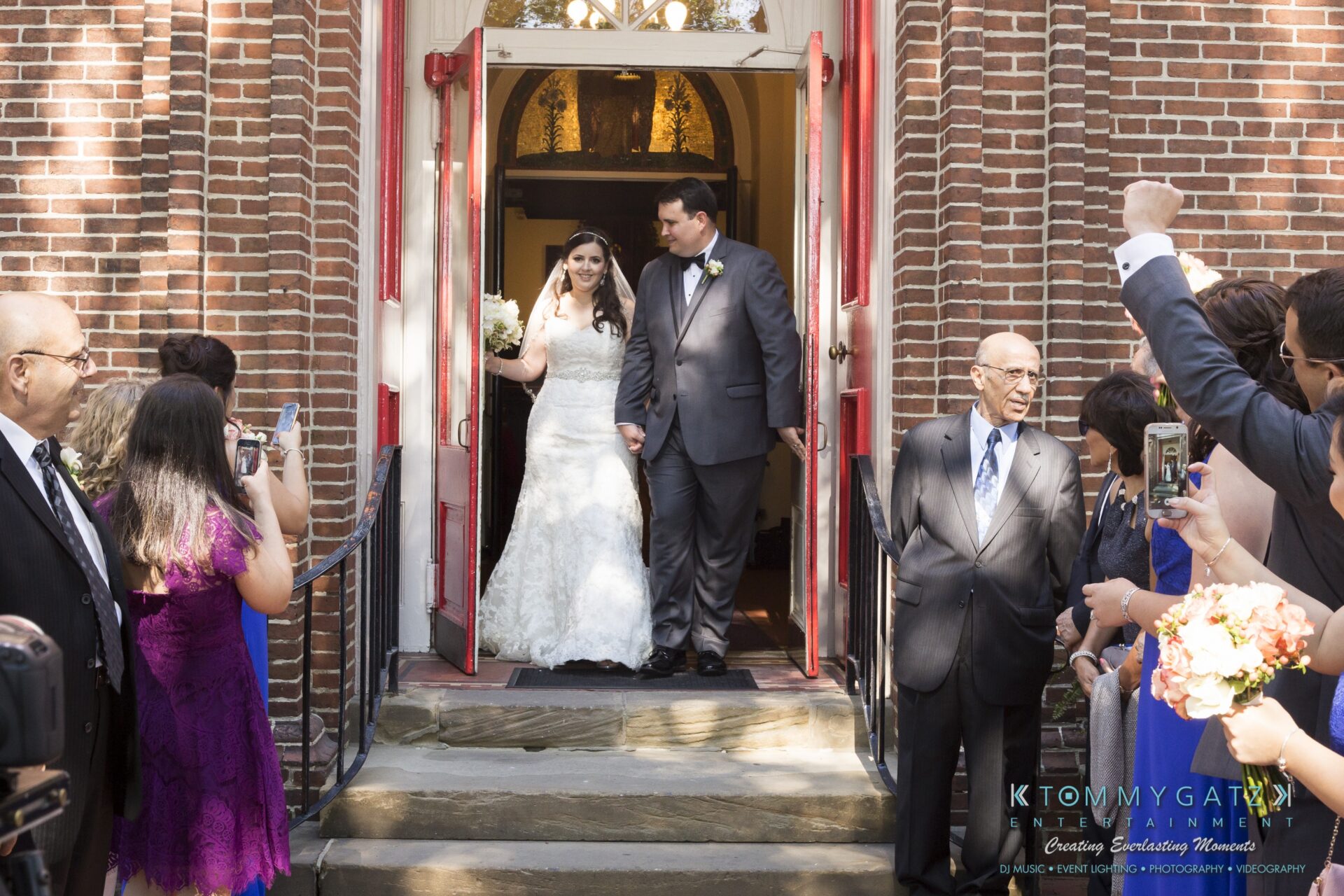 Jamie-and-Mark-Low-Res-Watermark-448 - TOMMY GATZ ENTERTAINMENT A bride and groom exit a brick building, holding hands. Guests stand around, some taking photos.