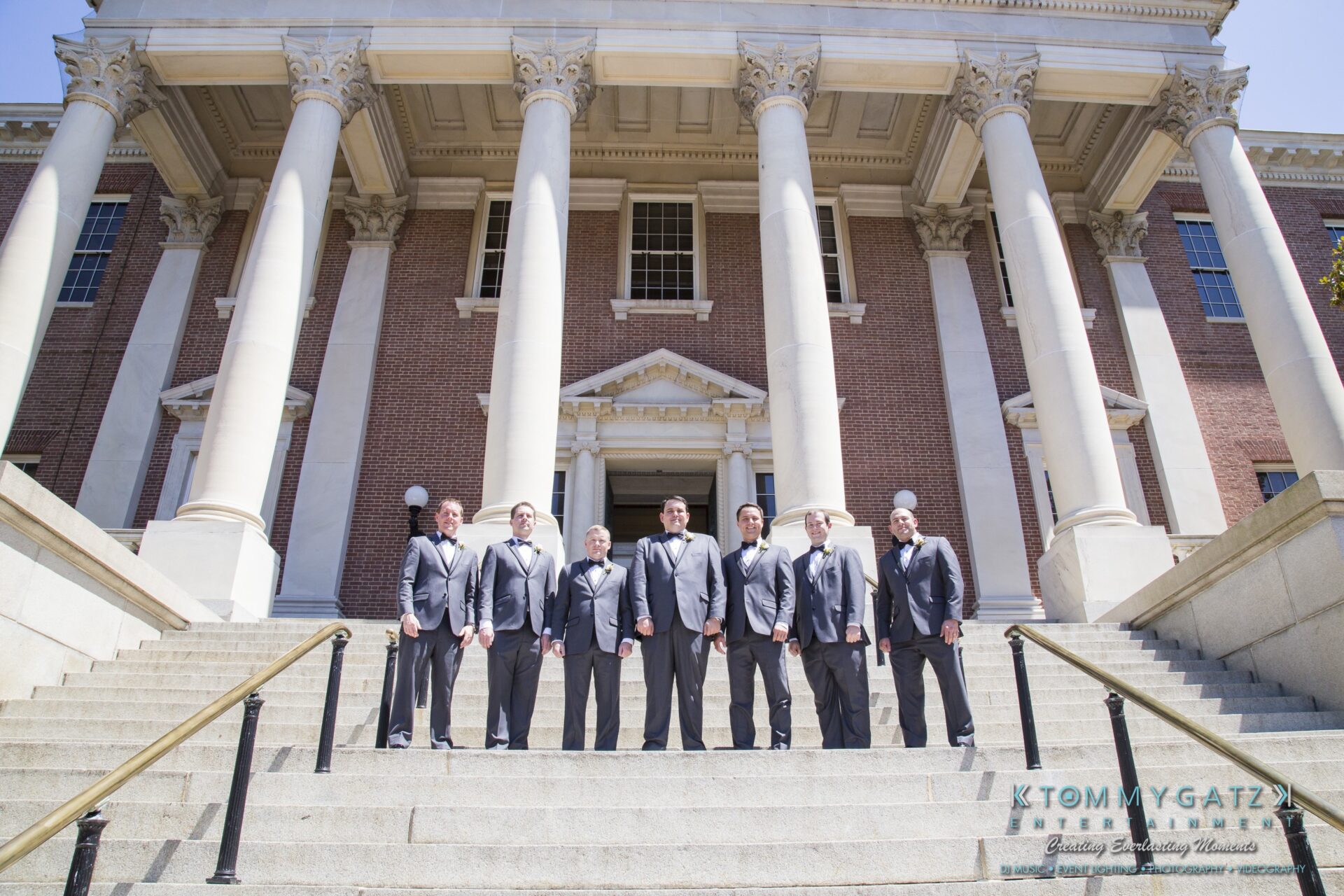 Jamie-and-Mark-Low-Res-Watermark-45 - TOMMY GATZ ENTERTAINMENT Seven people in suits stand on the steps of a large building with columns.