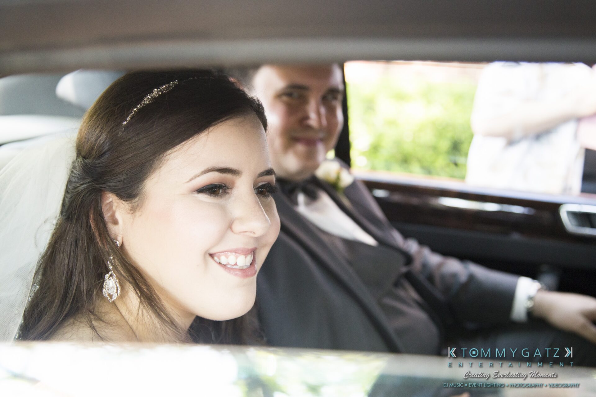 Jamie-and-Mark-Low-Res-Watermark-460 - TOMMY GATZ ENTERTAINMENT A bride and groom sit in the backseat of a car. The bride is smiling and wearing a veil and earrings. The groom is next to her, also smiling.