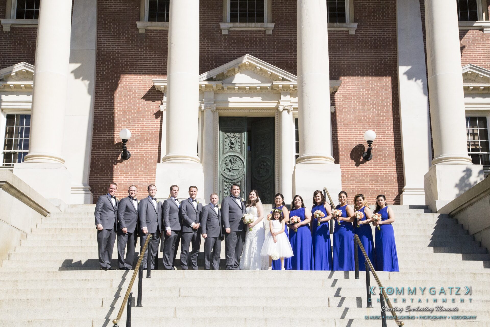 Jamie-and-Mark-Low-Res-Watermark-462 - TOMMY GATZ ENTERTAINMENT A wedding party stands on the steps of a brick building with columns. The groom and groomsmen wear gray suits, while the bride and bridesmaids wear blue dresses.