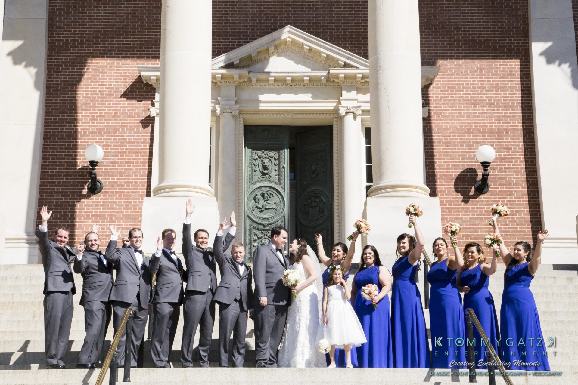 Jamie-and-Mark-Low-Res-Watermark-471 - TOMMY GATZ ENTERTAINMENT A wedding party dressed in gray suits and blue dresses poses with bouquets on the steps of a brick building with large columns.