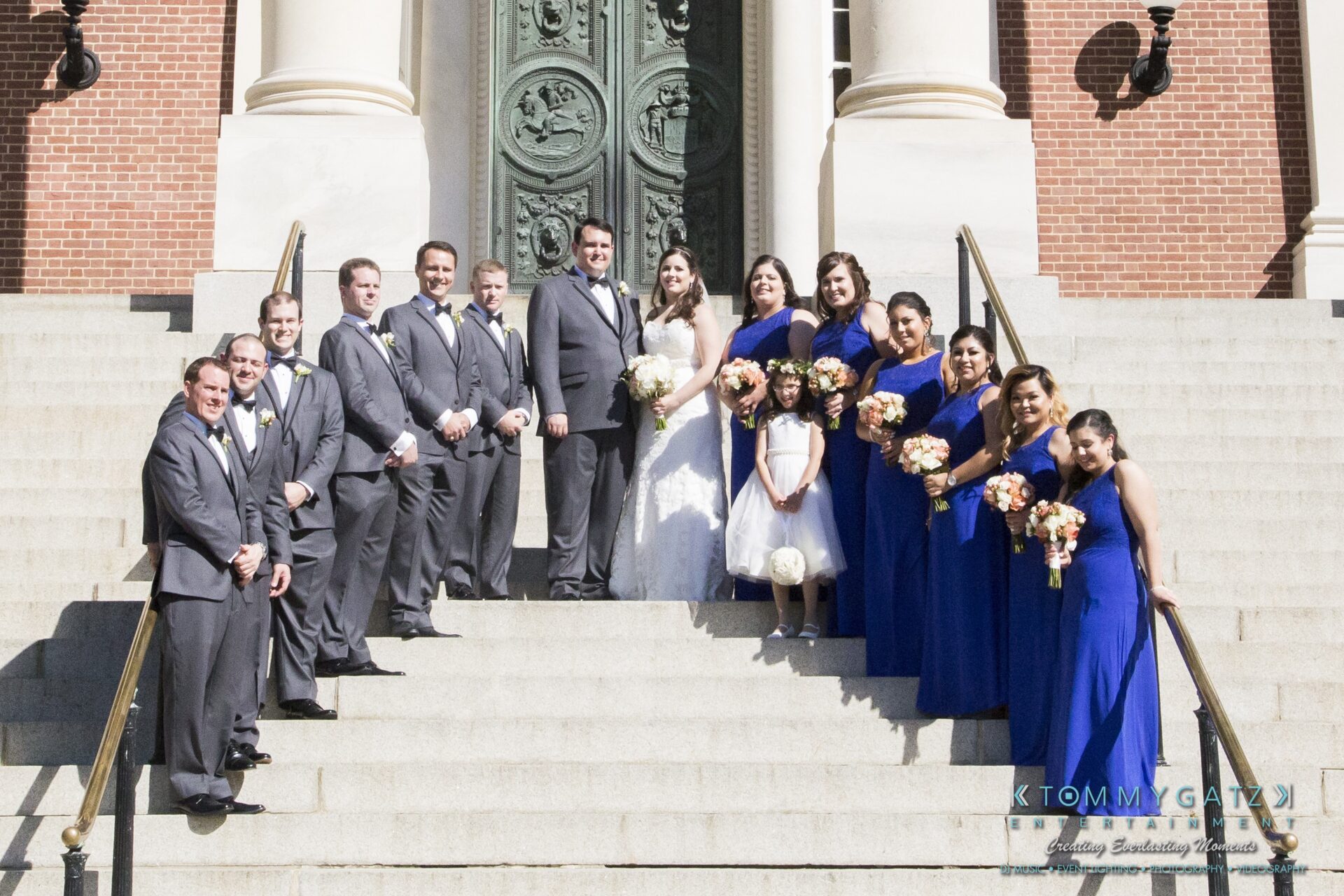 Jamie-and-Mark-Low-Res-Watermark-473 - TOMMY GATZ ENTERTAINMENT A wedding party stands on steps. The group includes a bride, groom, and bridesmaids in blue dresses, along with groomsmen in gray suits, posed in front of a large ornate door.