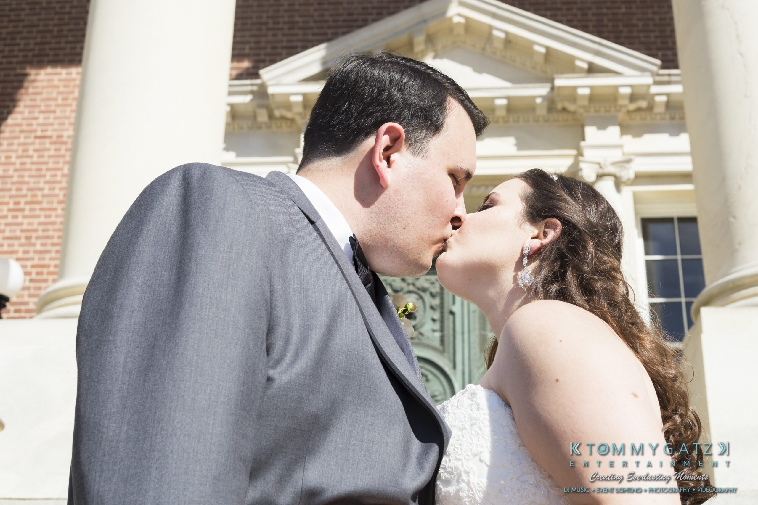 Jamie-and-Mark-Low-Res-Watermark-486 - TOMMY GATZ ENTERTAINMENT A couple dressed in wedding attire shares a kiss in front of a building with columns.