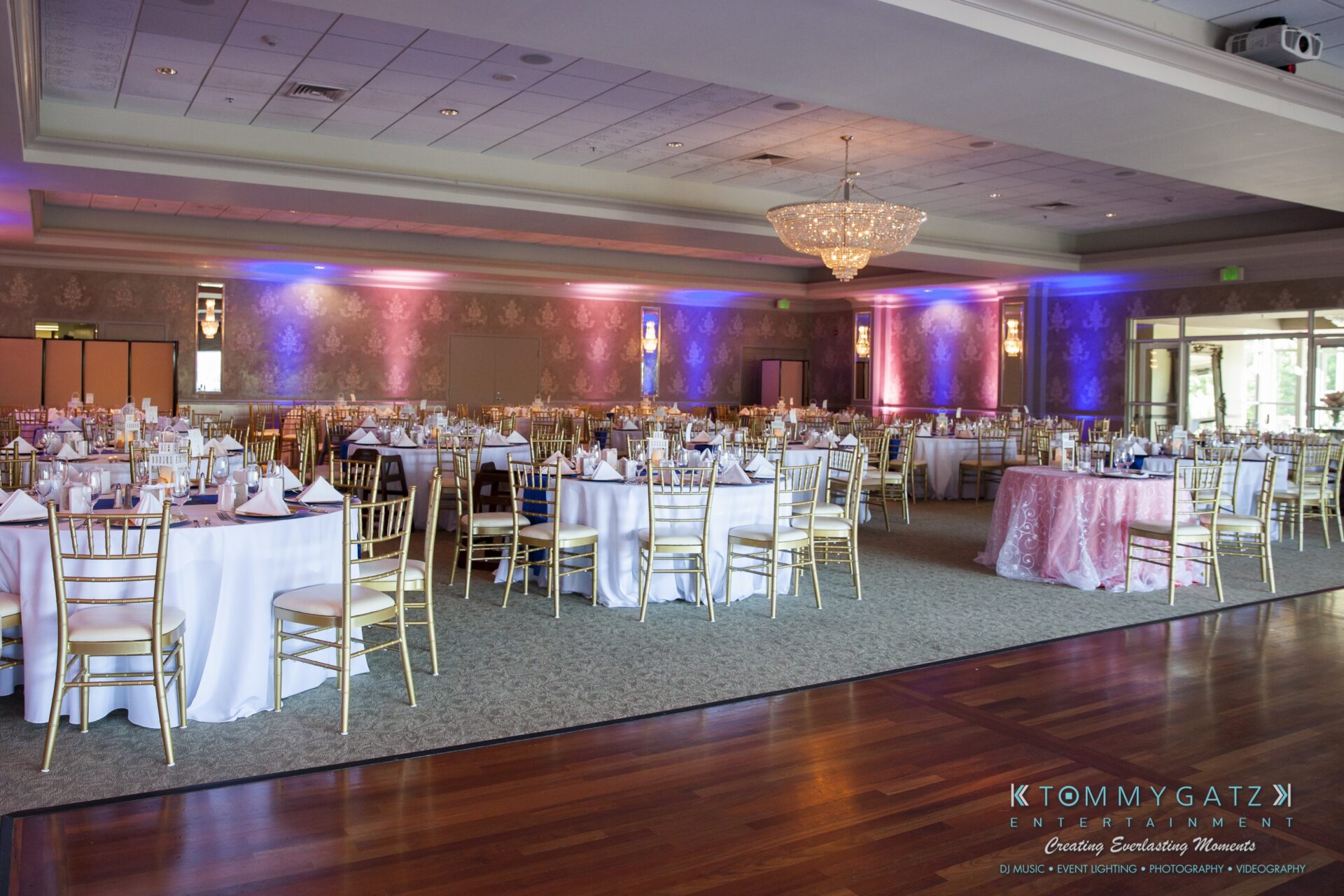 Jamie-and-Mark-Low-Res-Watermark-487 - TOMMY GATZ ENTERTAINMENT Spacious banquet hall set up for an event with round tables, white tablecloths, gold chairs, place settings, and elegant lighting.