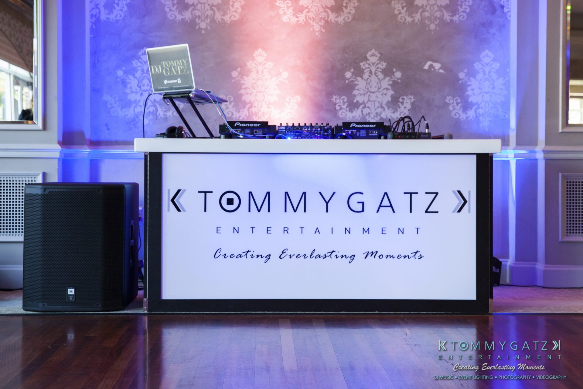 Jamie-and-Mark-Low-Res-Watermark-489 - TOMMY GATZ ENTERTAINMENT DJ booth with branding signage reading "Tommy Gatz Entertainment" and "Creating Everlasting Moments." Equipment includes a laptop and audio gear. Venue features elegant lighting and decor.