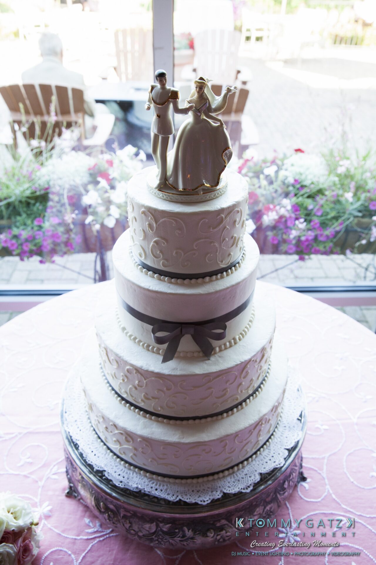 Jamie-and-Mark-Low-Res-Watermark-491 - TOMMY GATZ ENTERTAINMENT A three-tiered wedding cake with white icing and decorative patterns, topped with a figurine of a couple dancing.