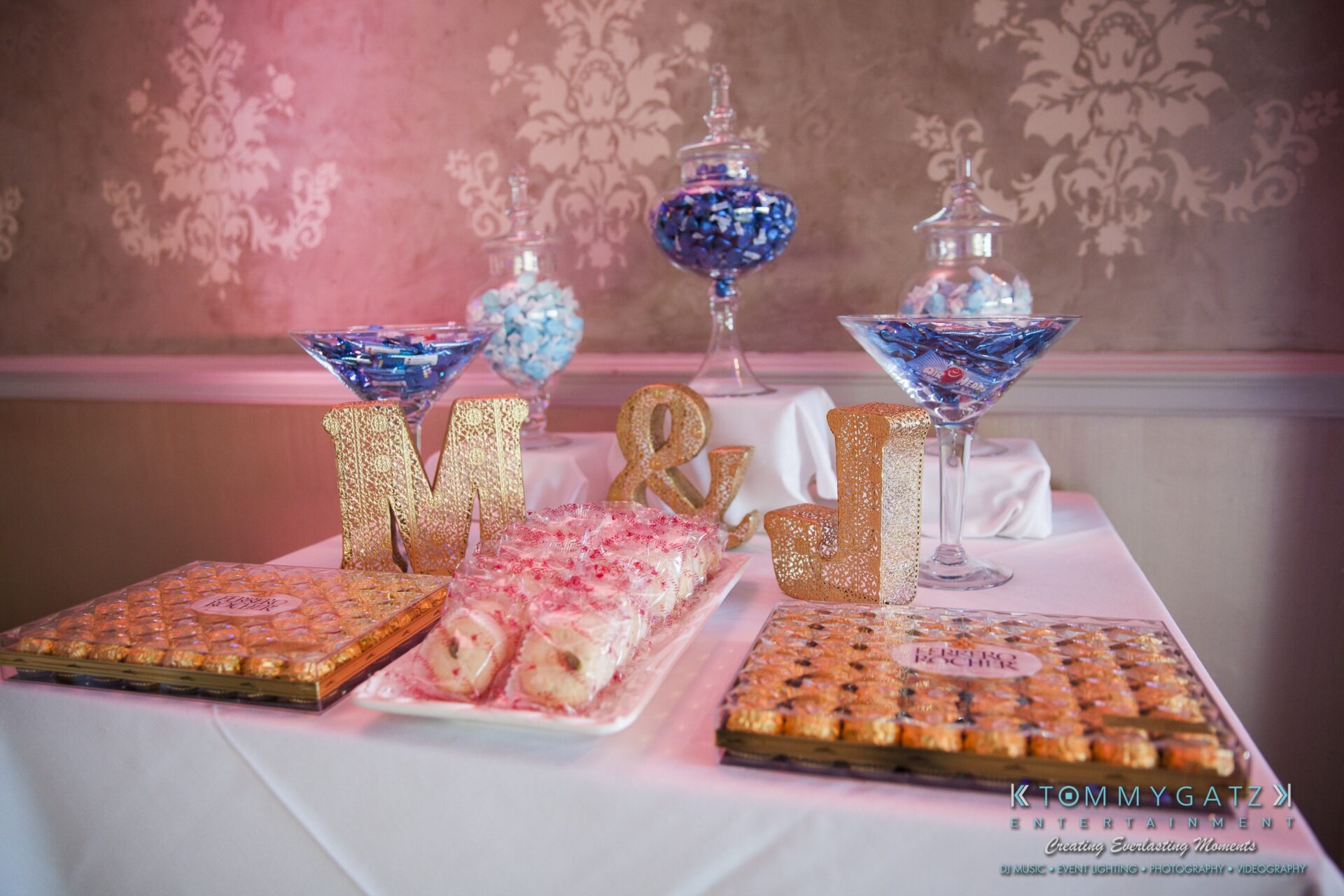 Jamie-and-Mark-Low-Res-Watermark-494 - TOMMY GATZ ENTERTAINMENT Table with gold glitter letters "M" and "J", two trays of pastries, and three large glasses filled with blue and white candies.