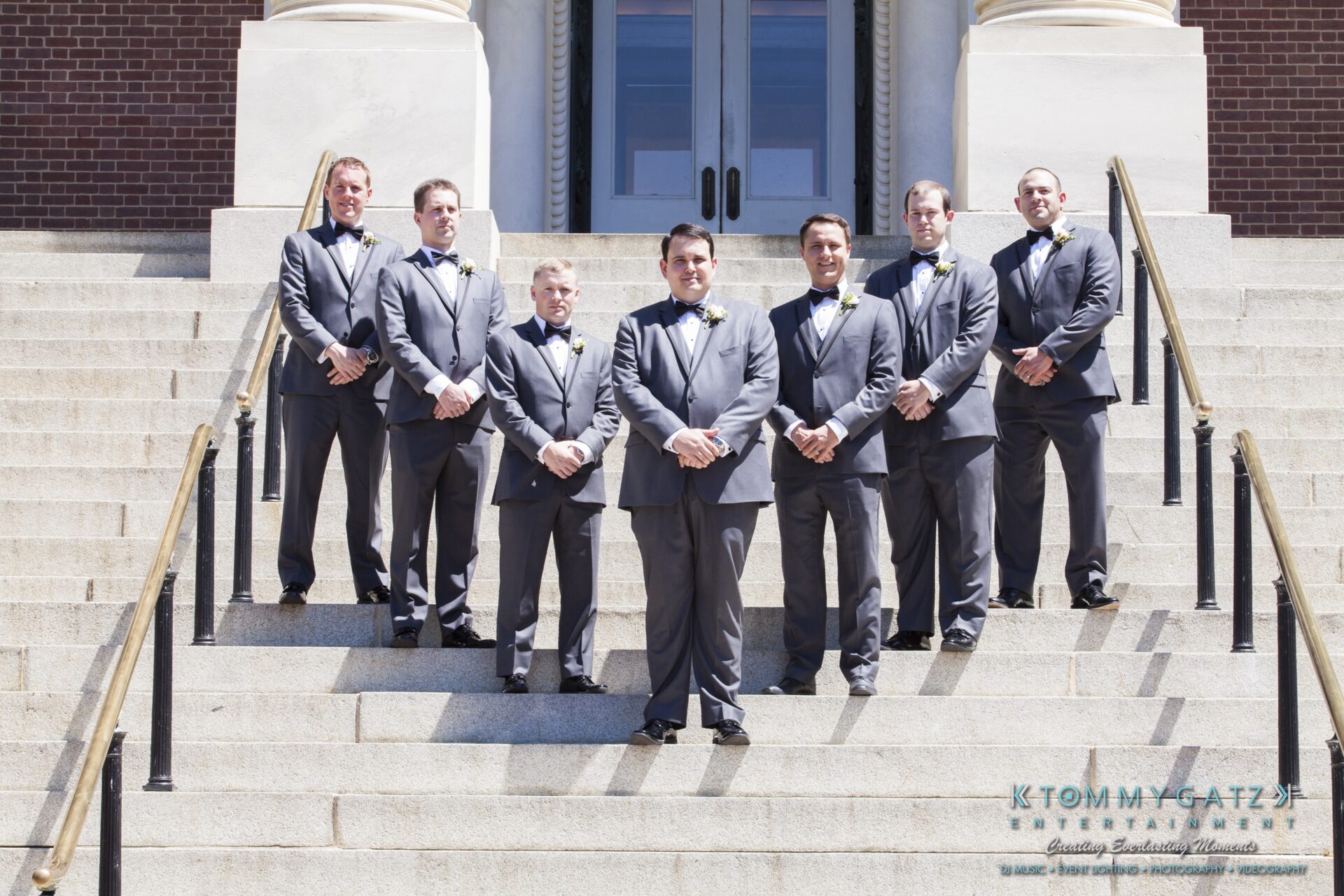 Jamie-and-Mark-Low-Res-Watermark-50 - TOMMY GATZ ENTERTAINMENT Seven men in gray suits with bow ties stand on steps in front of a building with large doors.