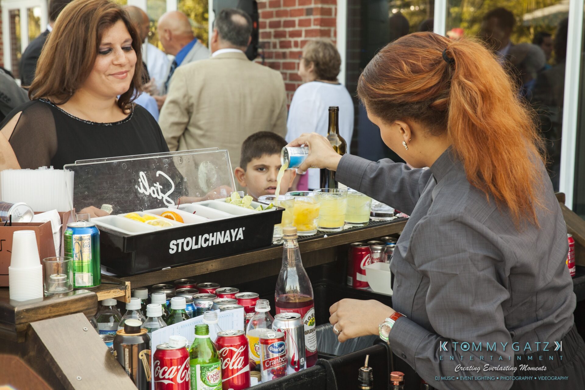 Jamie-and-Mark-Low-Res-Watermark-505 - TOMMY GATZ ENTERTAINMENT A bartender prepares drinks at an outdoor event, pouring a can of Red Bull into a glass. A woman and a child stand at the bar. Various soft drinks are visible on the counter.