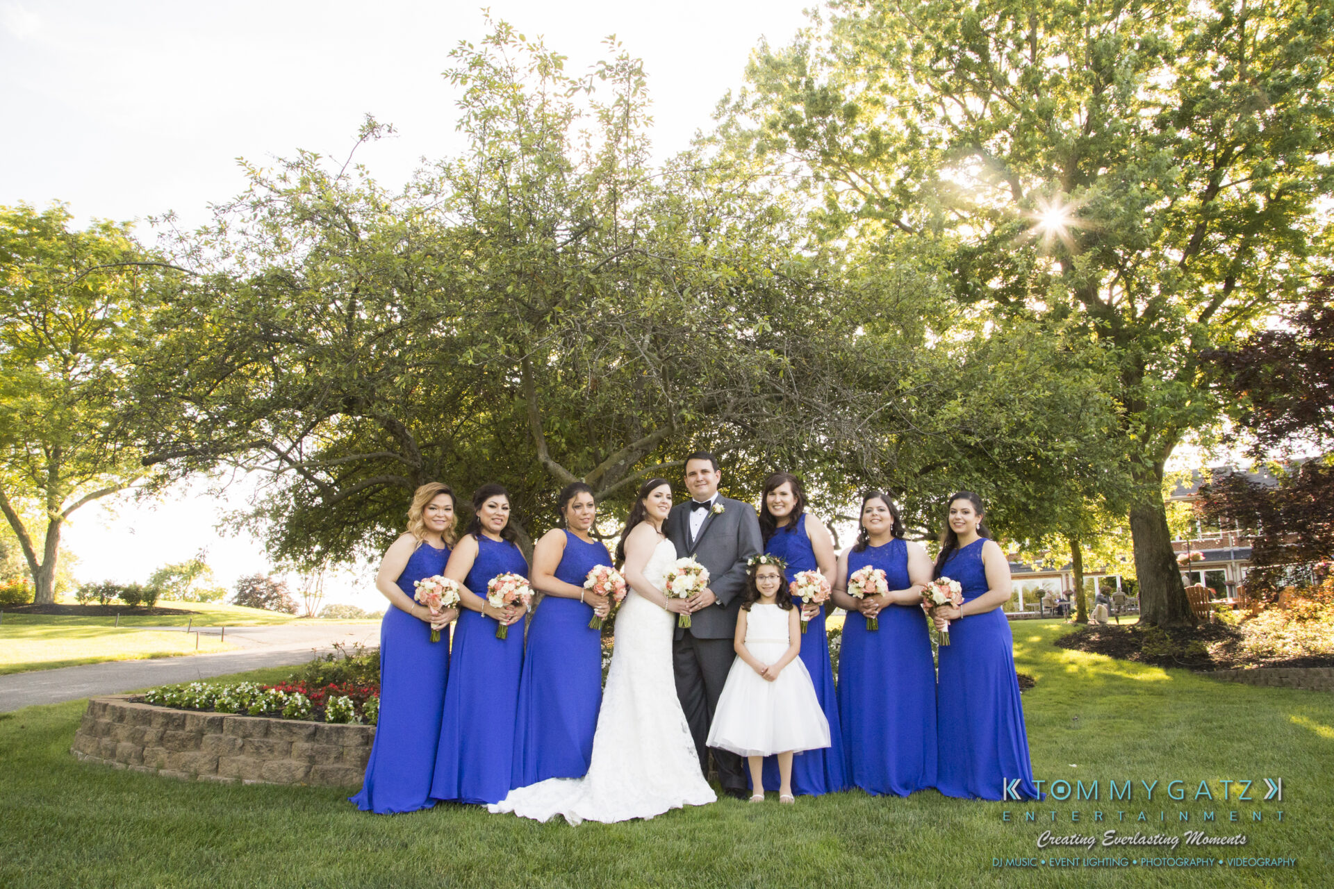 Jamie-and-Mark-Low-Res-Watermark-519 - TOMMY GATZ ENTERTAINMENT A bride in a white gown stands with bridesmaids in blue dresses, holding bouquets. A groom and a flower girl are also present, with a garden and tree in the background.