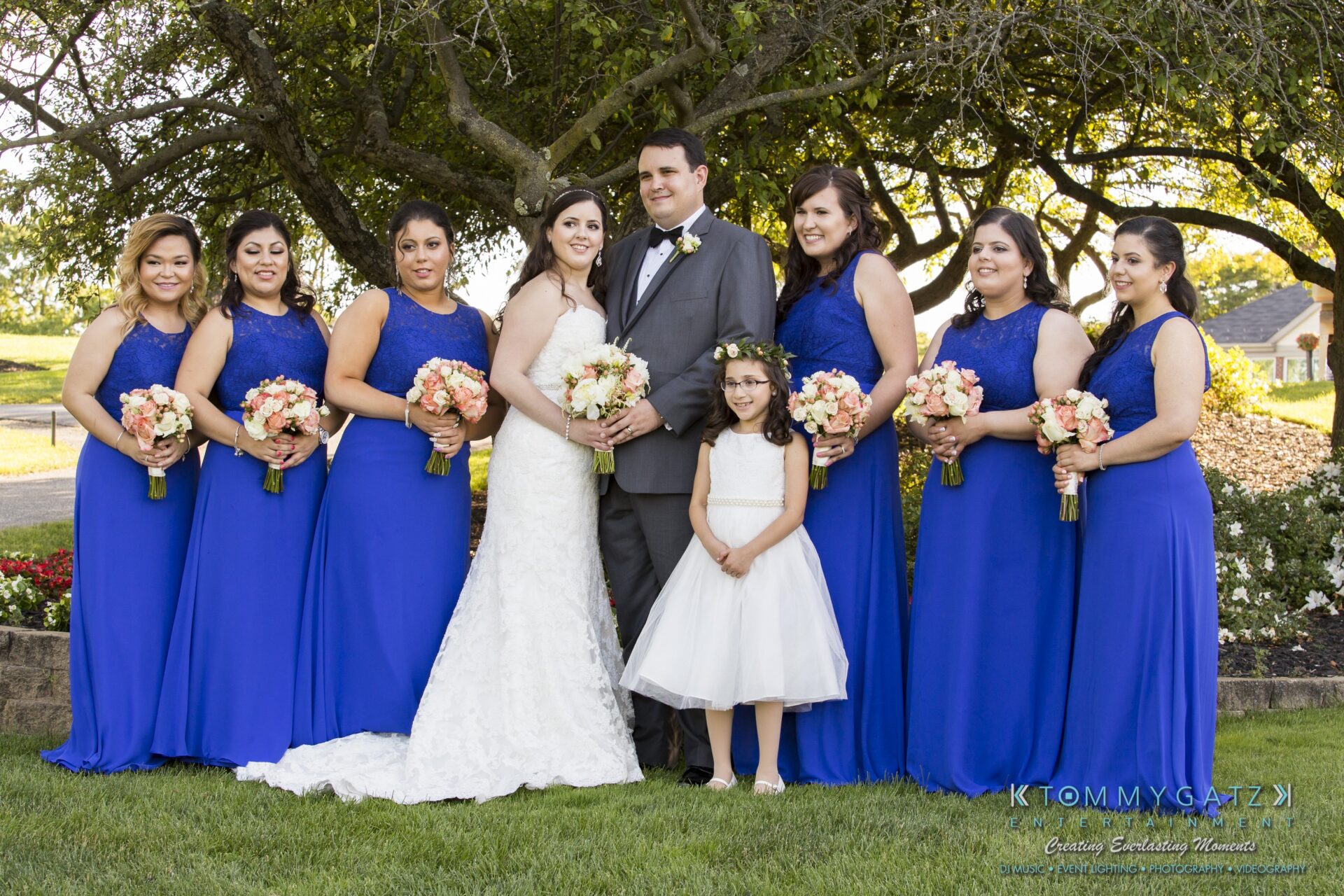 Jamie-and-Mark-Low-Res-Watermark-522 - TOMMY GATZ ENTERTAINMENT A bride and groom pose with a flower girl and six bridesmaids wearing blue dresses, standing outdoors in front of a tree.