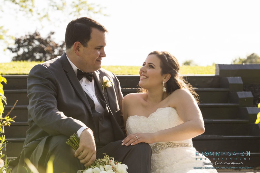 Jamie-and-Mark-Low-Res-Watermark-568-1024x683 - TOMMY GATZ ENTERTAINMENT A bride and groom sit together on outdoor steps, both smiling. The groom wears a dark suit, and the bride wears a white strapless dress, holding a bouquet.