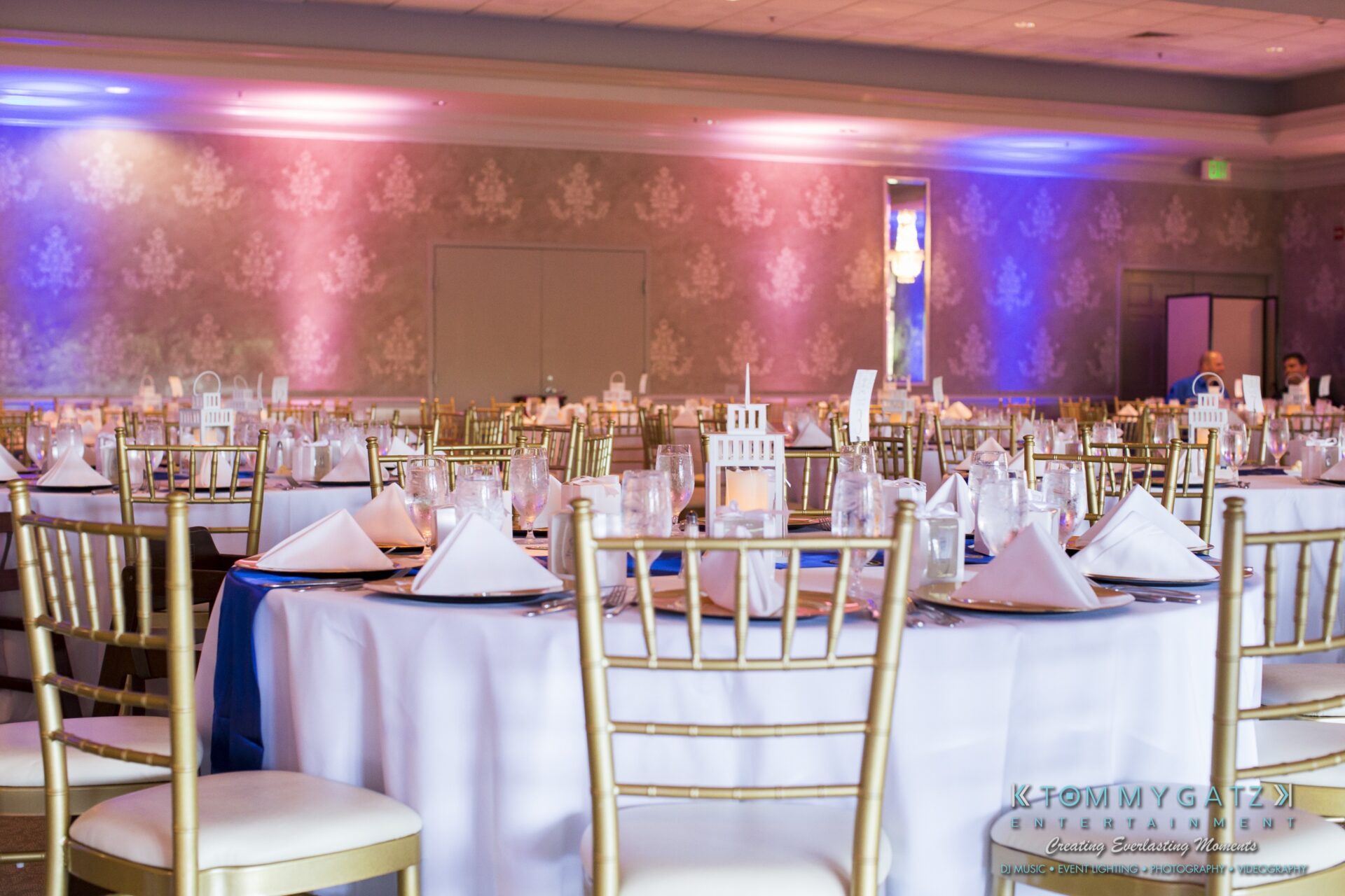 Jamie-and-Mark-Low-Res-Watermark-583 - TOMMY GATZ ENTERTAINMENT Banquet hall set for an event with round tables, white tablecloths, folded napkins, gold chairs, and ambient lighting in blue and purple tones.
