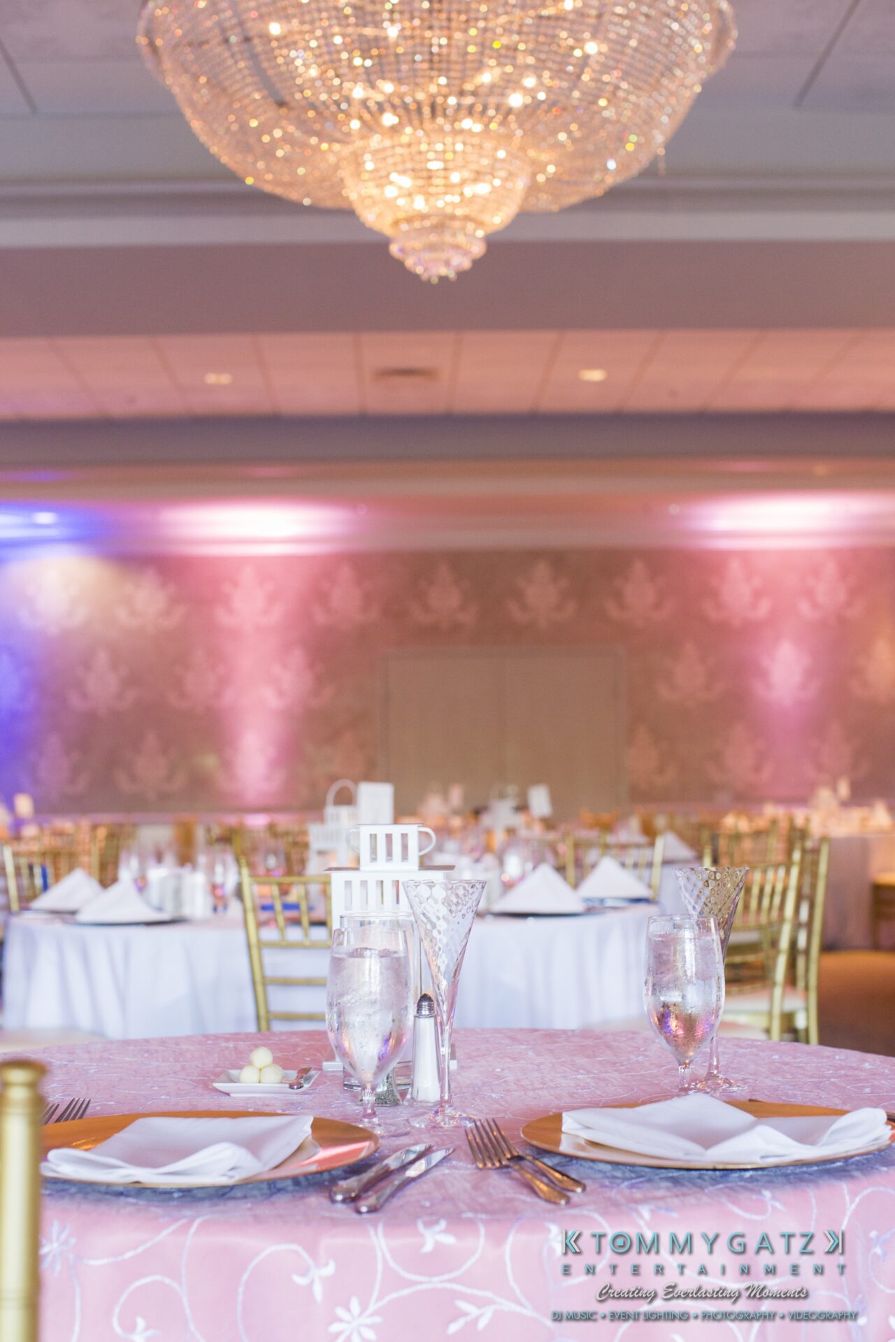 Jamie-and-Mark-Low-Res-Watermark-585-1 - TOMMY GATZ ENTERTAINMENT Elegant banquet hall with chandeliers, set with round tables, gold chairs, and crystal glasses. Tables are adorned with pink tablecloths and folded napkins. Soft lighting in background.