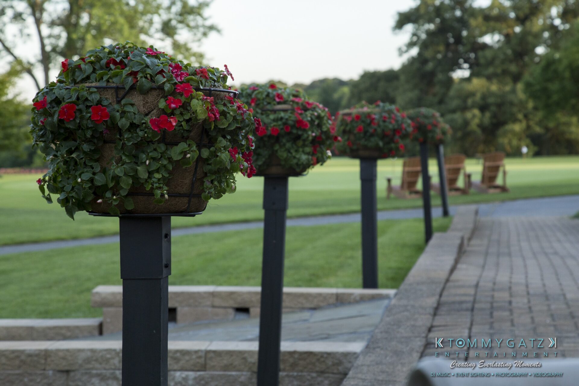 Jamie-and-Mark-Low-Res-Watermark-635 - TOMMY GATZ ENTERTAINMENT A row of flower baskets with red blooms on black posts lines a stone path with green grass and trees in the background.