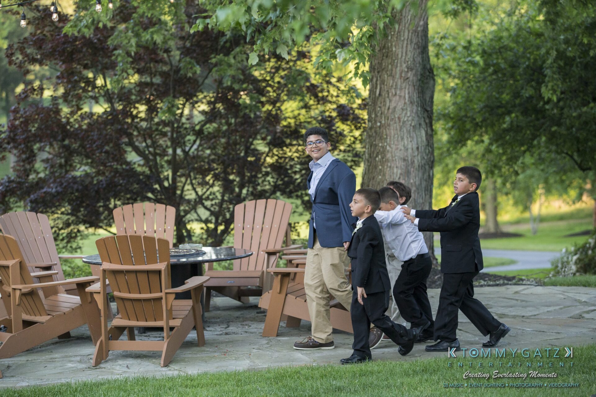 Jamie-and-Mark-Low-Res-Watermark-638 - TOMMY GATZ ENTERTAINMENT Four young boys in suits walk in a line, following an adult male in casual clothing, near wooden chairs and a tree in a garden setting.