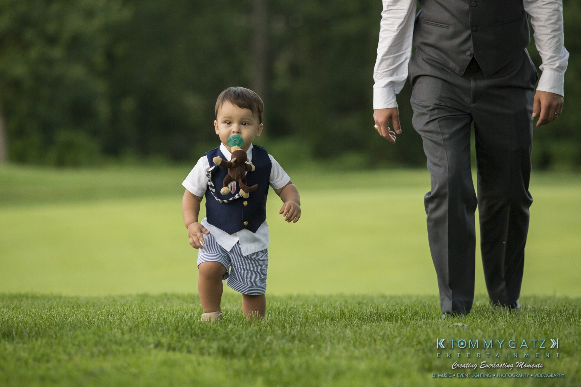 Jamie-and-Mark-Low-Res-Watermark-640 - TOMMY GATZ ENTERTAINMENT A toddler with a pacifier and stuffed toy walks on a grassy field, followed by a man in a vest and tie.