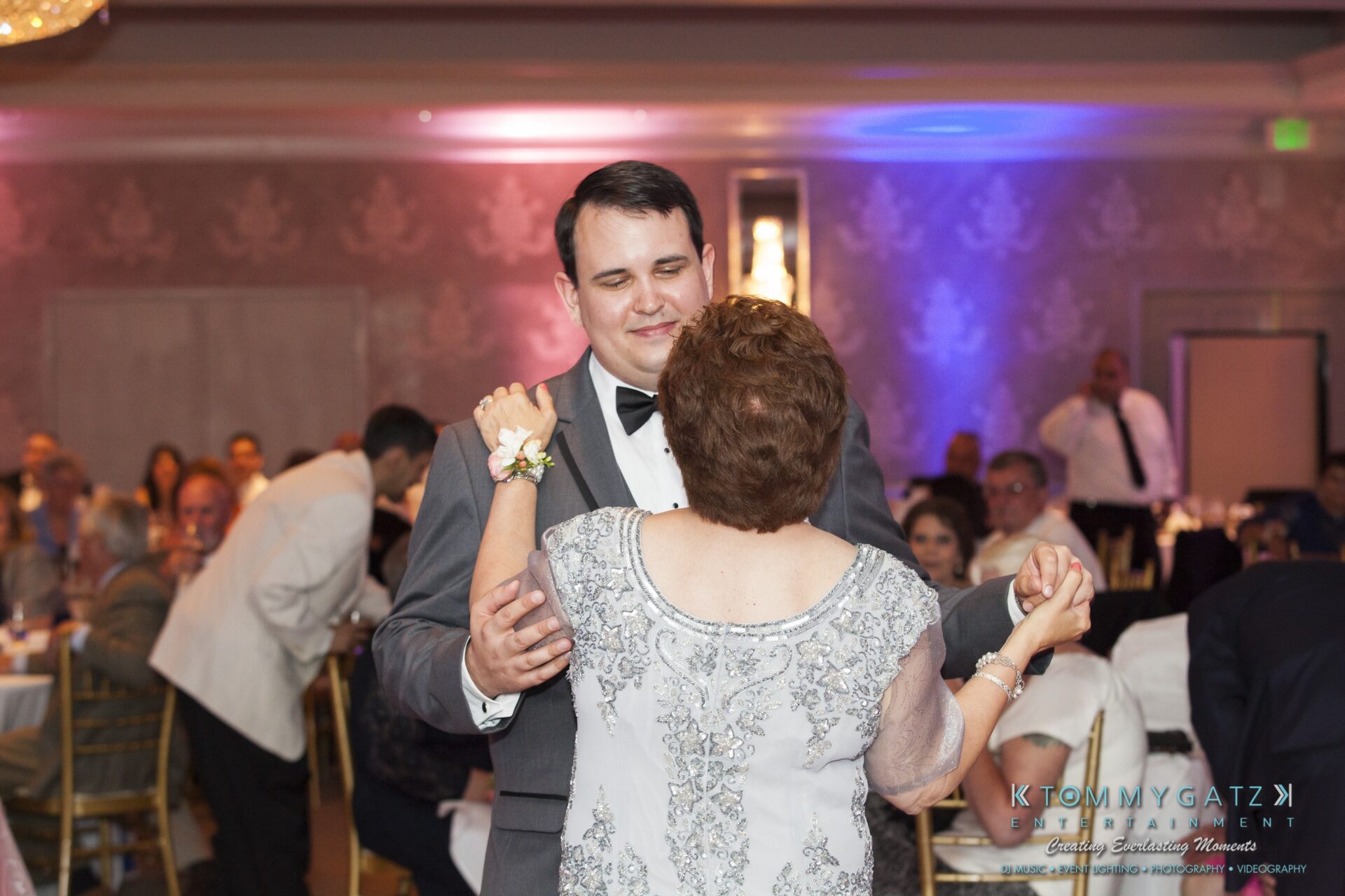 Jamie-and-Mark-Low-Res-Watermark-675 - TOMMY GATZ ENTERTAINMENT A groom and an older woman dance together in a formal setting, with guests seated in the background.