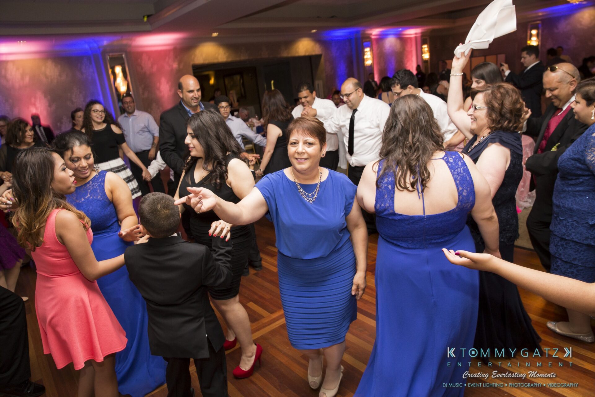 Jamie-and-Mark-Low-Res-Watermark-693 - TOMMY GATZ ENTERTAINMENT A group of people dancing at a formal event in a decorated venue, with colorful lighting and a wooden floor.
