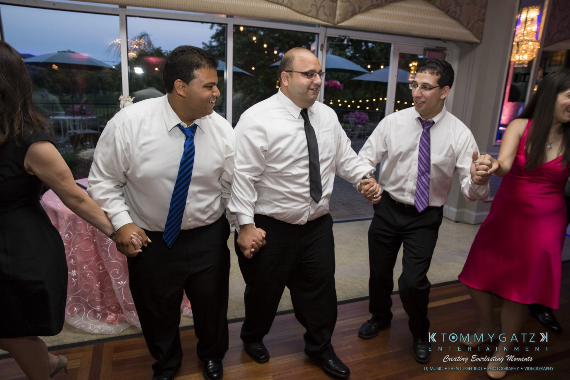 Jamie-and-Mark-Low-Res-Watermark-694 - TOMMY GATZ ENTERTAINMENT A group of people in formal attire hold hands in a dance circle at an indoor event.