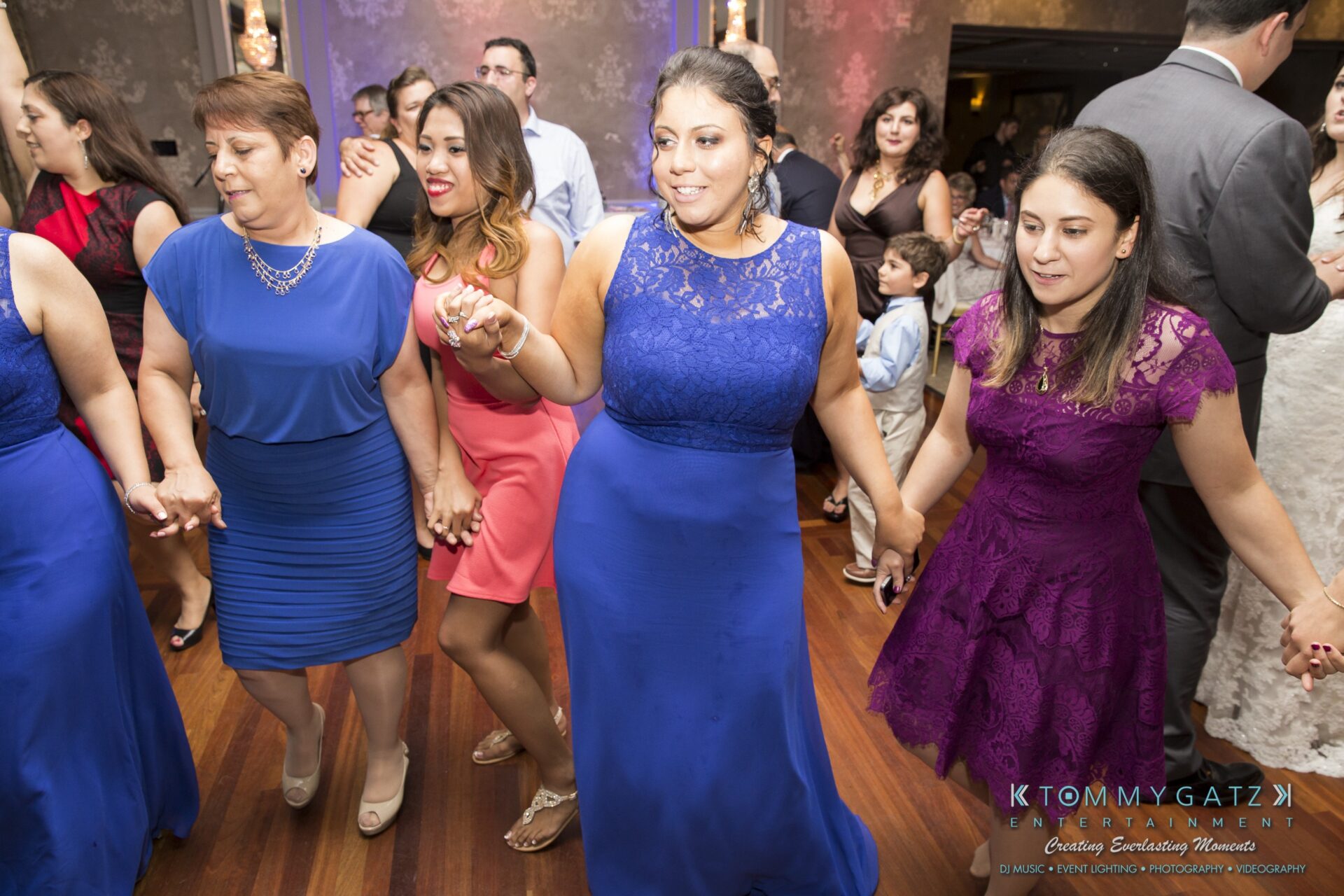 Jamie-and-Mark-Low-Res-Watermark-696 - TOMMY GATZ ENTERTAINMENT A group of people in colorful dresses holding hands and dancing at a formal event in a decorated indoor venue.