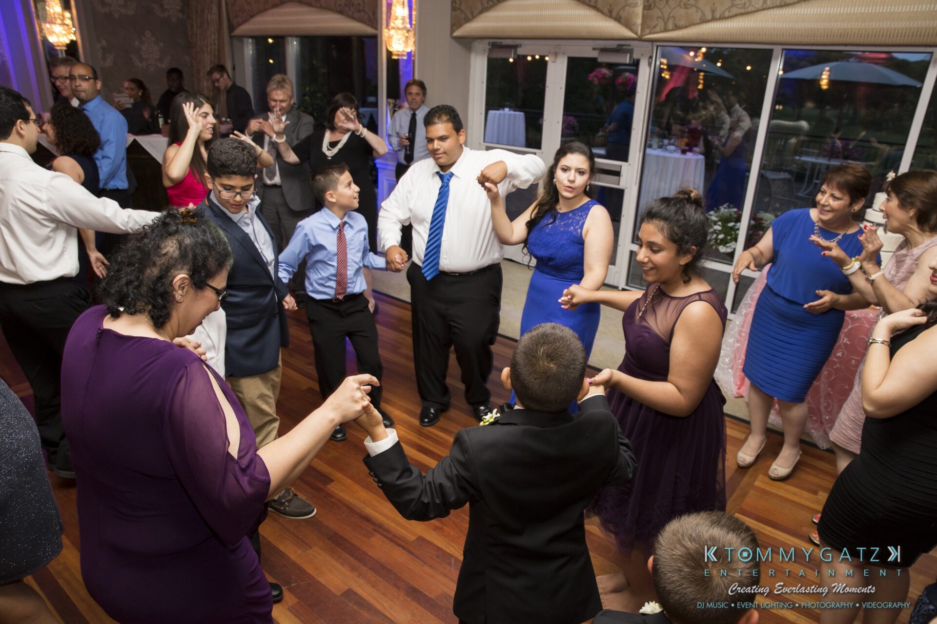 Jamie-and-Mark-Low-Res-Watermark-703 - TOMMY GATZ ENTERTAINMENT A group of people, including adults and children, dance together on a wooden floor in a dressed-up event setting.