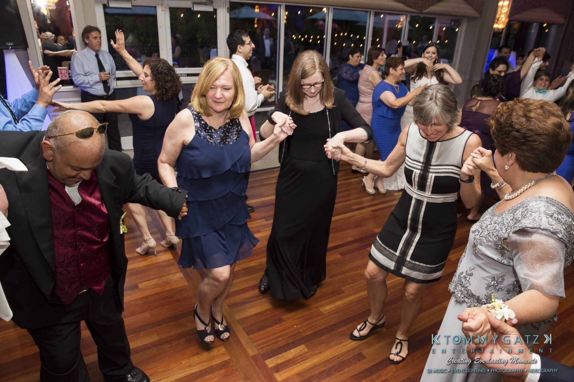 Jamie-and-Mark-Low-Res-Watermark-708 - TOMMY GATZ ENTERTAINMENT A group of people joyfully dancing, holding hands, on a wooden floor in a brightly lit event space.