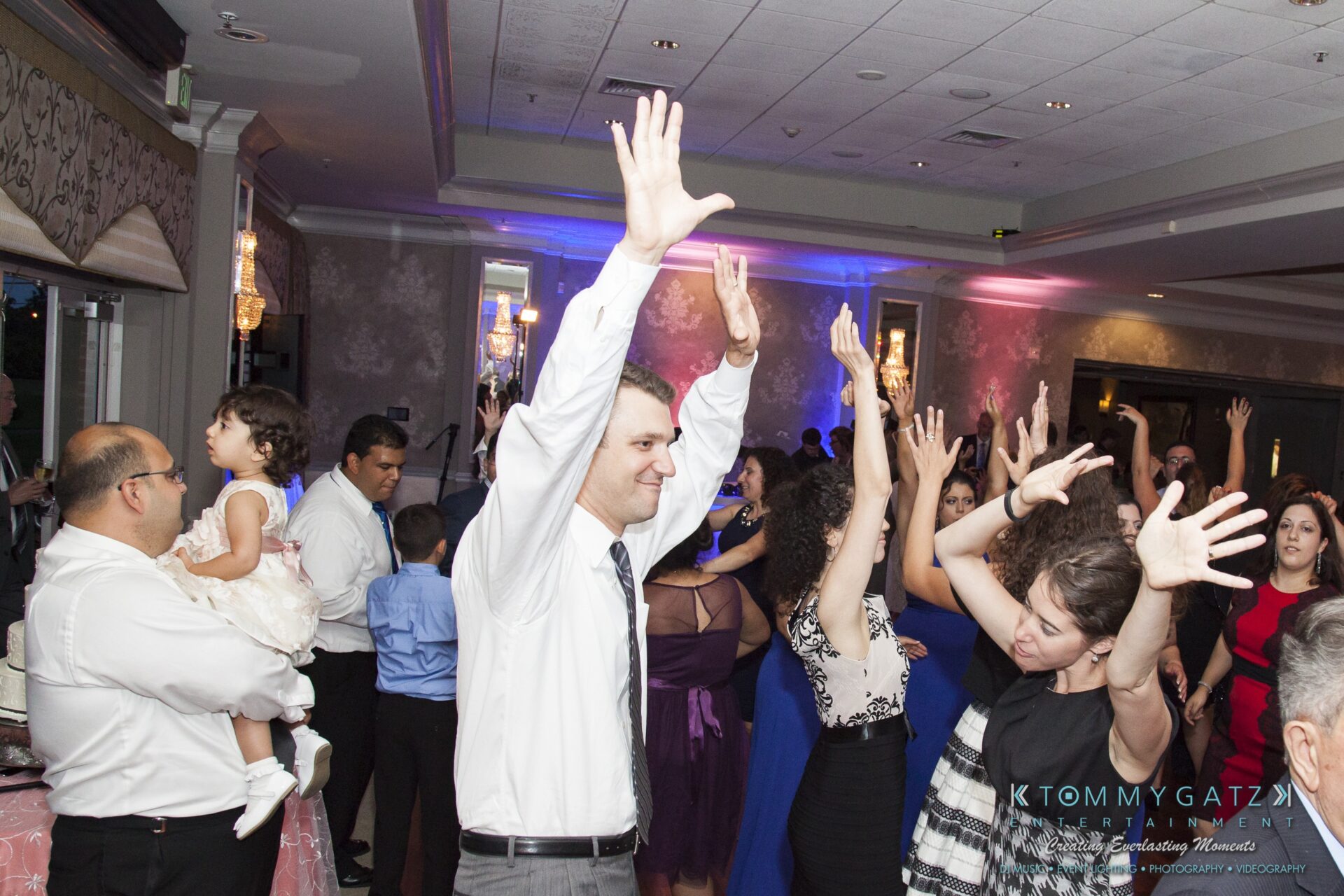 Jamie-and-Mark-Low-Res-Watermark-728 - TOMMY GATZ ENTERTAINMENT People are dancing with their hands raised at an indoor event. Some are dressed in formal attire.