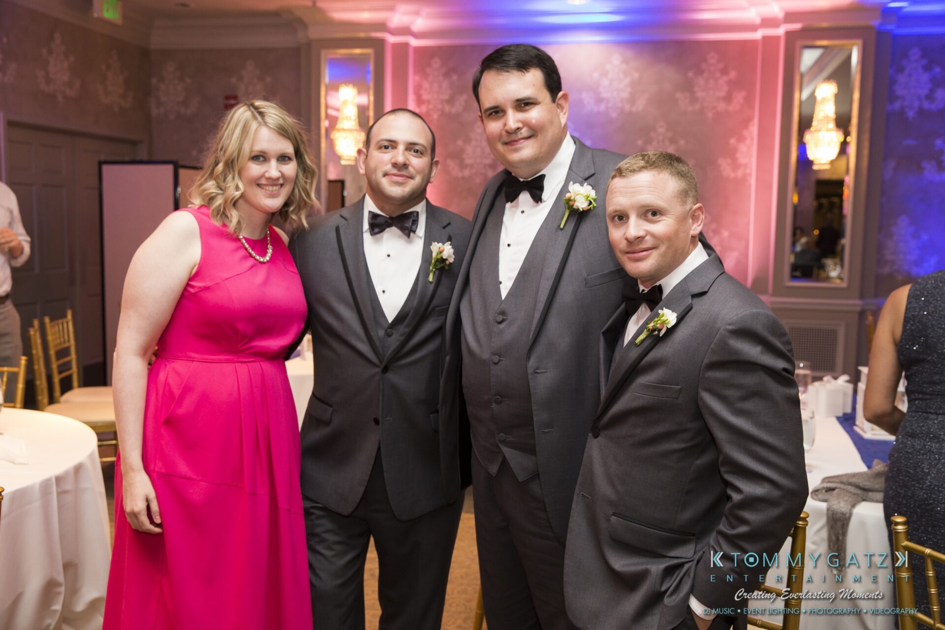Jamie-and-Mark-Low-Res-Watermark-736 - TOMMY GATZ ENTERTAINMENT Four people, three men in suits and one woman in a pink dress, pose together at a formal event in a decorated room.