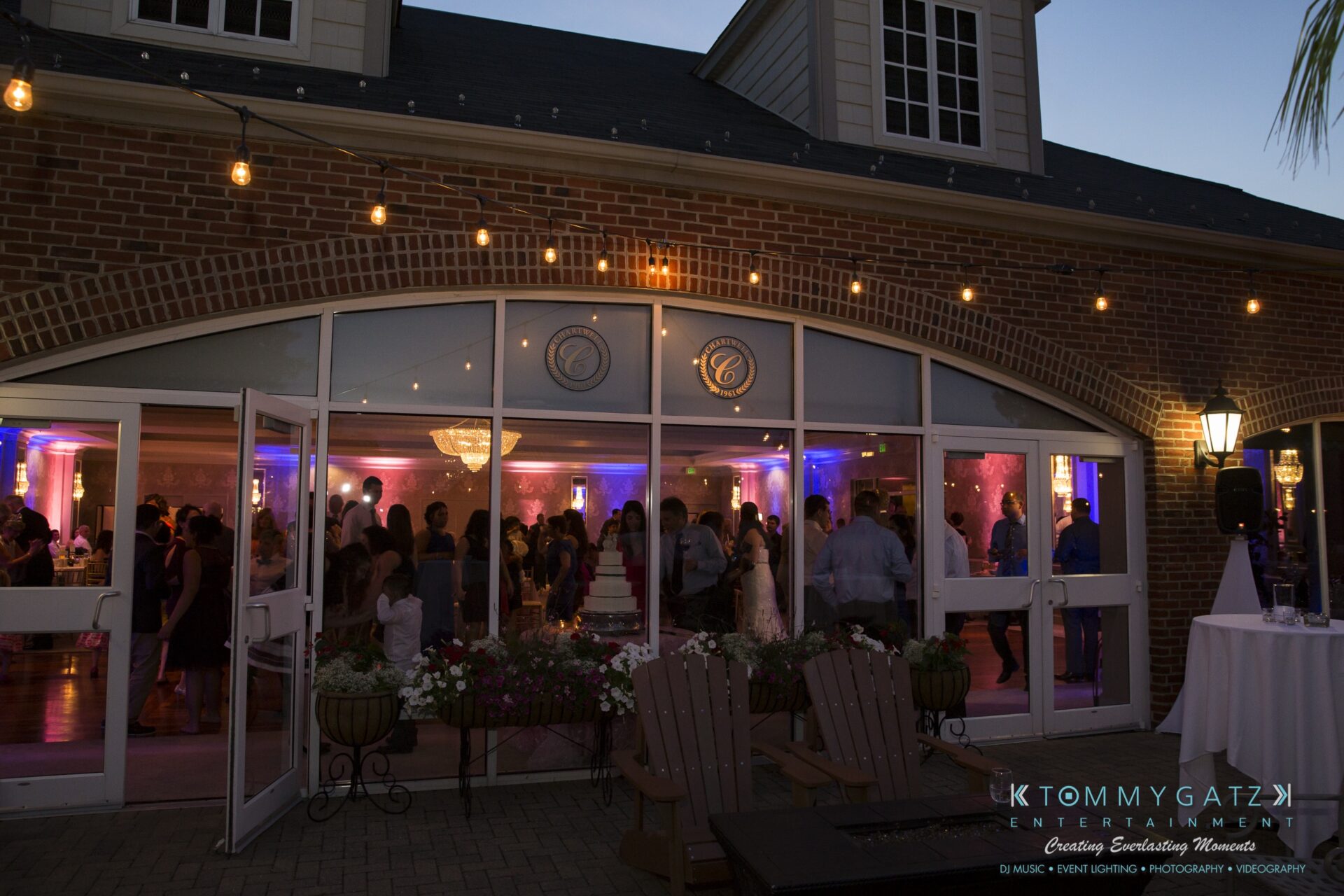 Jamie-and-Mark-Low-Res-Watermark-739 - TOMMY GATZ ENTERTAINMENT People at a lit indoor party venue with large windows, brick walls, and string lights outside.