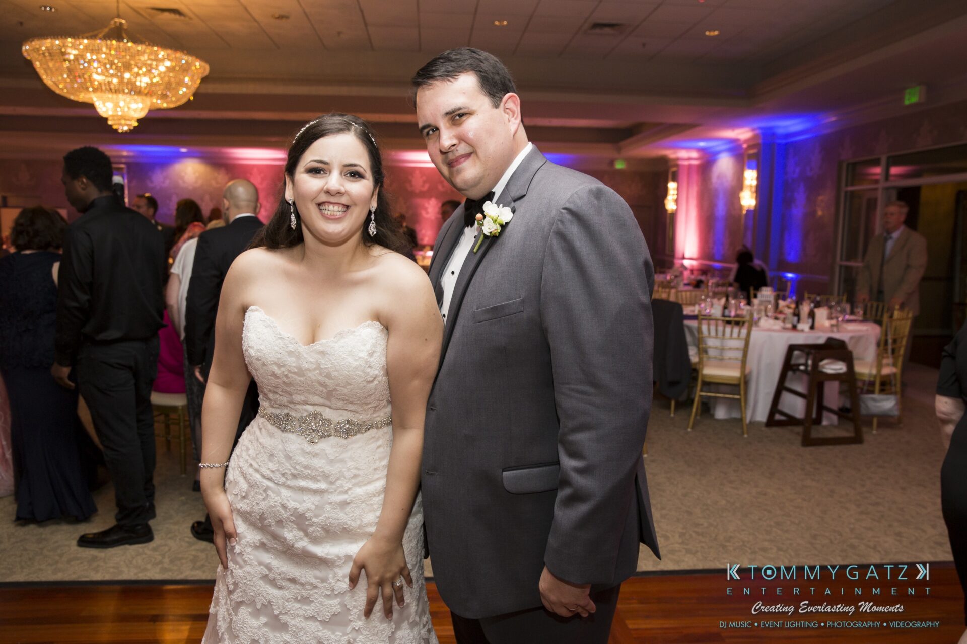 Jamie-and-Mark-Low-Res-Watermark-745 - TOMMY GATZ ENTERTAINMENT A bride and groom smile at their wedding reception, with guests in the background and a decorative chandelier overhead.