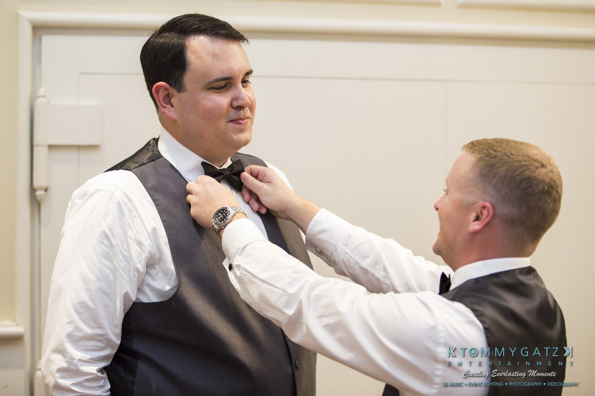 Jamie-and-Mark-Low-Res-Watermark-75 - TOMMY GATZ ENTERTAINMENT A man adjusts another man's bow tie in a formal setting. Both are wearing vests and dress shirts.
