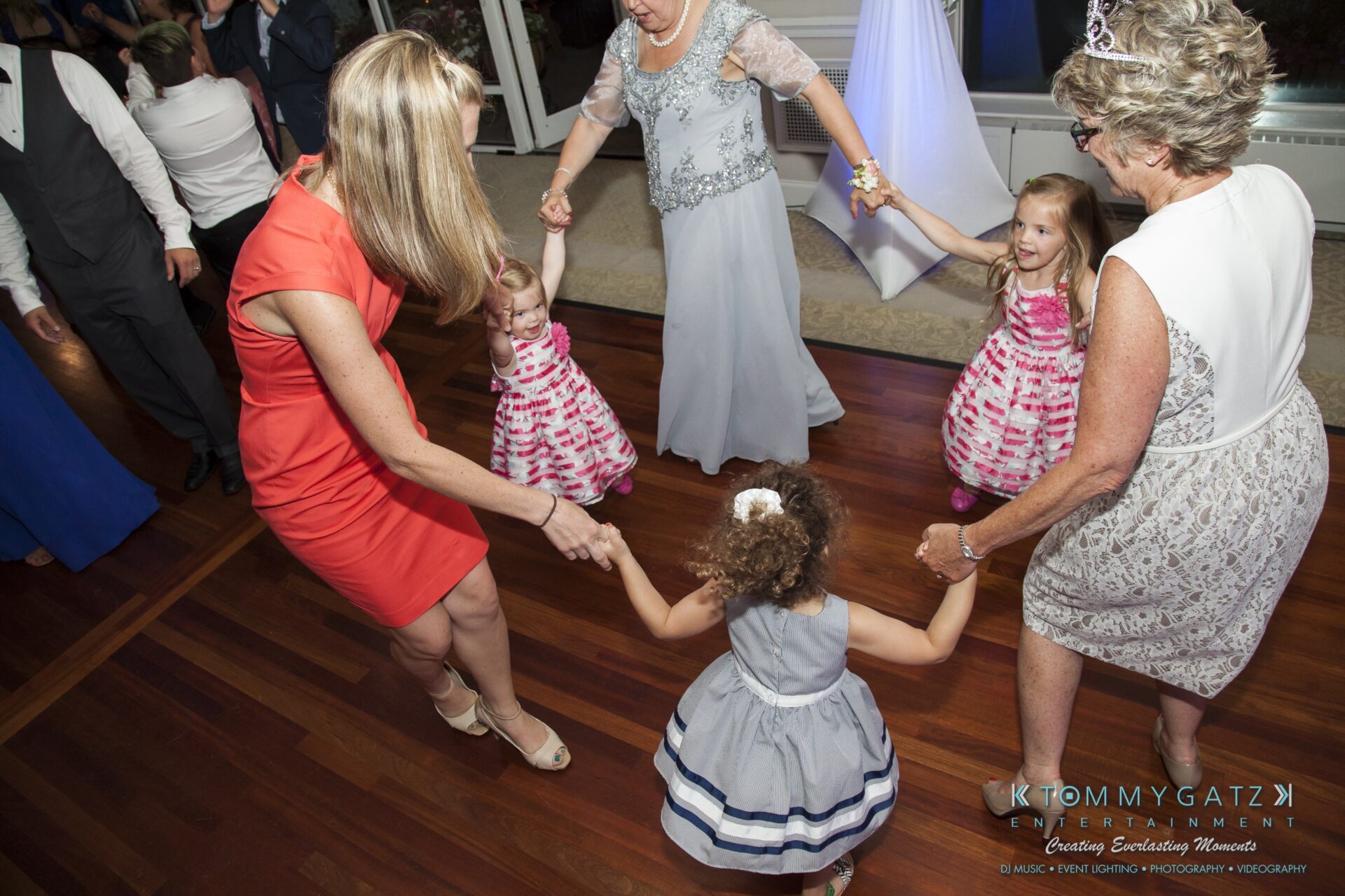 Jamie-and-Mark-Low-Res-Watermark-752 - TOMMY GATZ ENTERTAINMENT A group of people, including children in dresses, dance in a circle on a polished wooden floor at an indoor event.