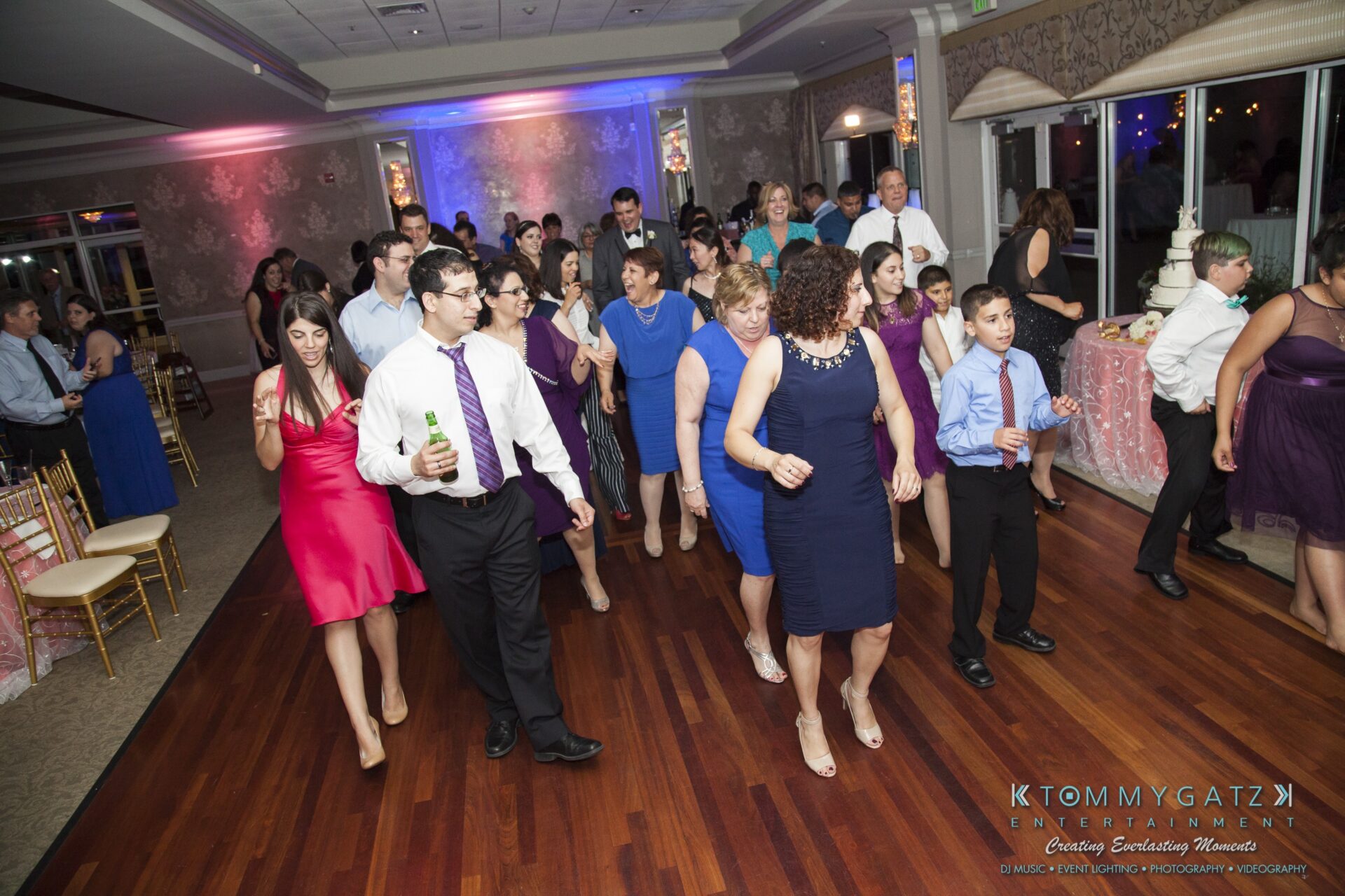 Jamie-and-Mark-Low-Res-Watermark-755 - TOMMY GATZ ENTERTAINMENT A group of people dance together in a decorated indoor venue, with colorful lights in the background.